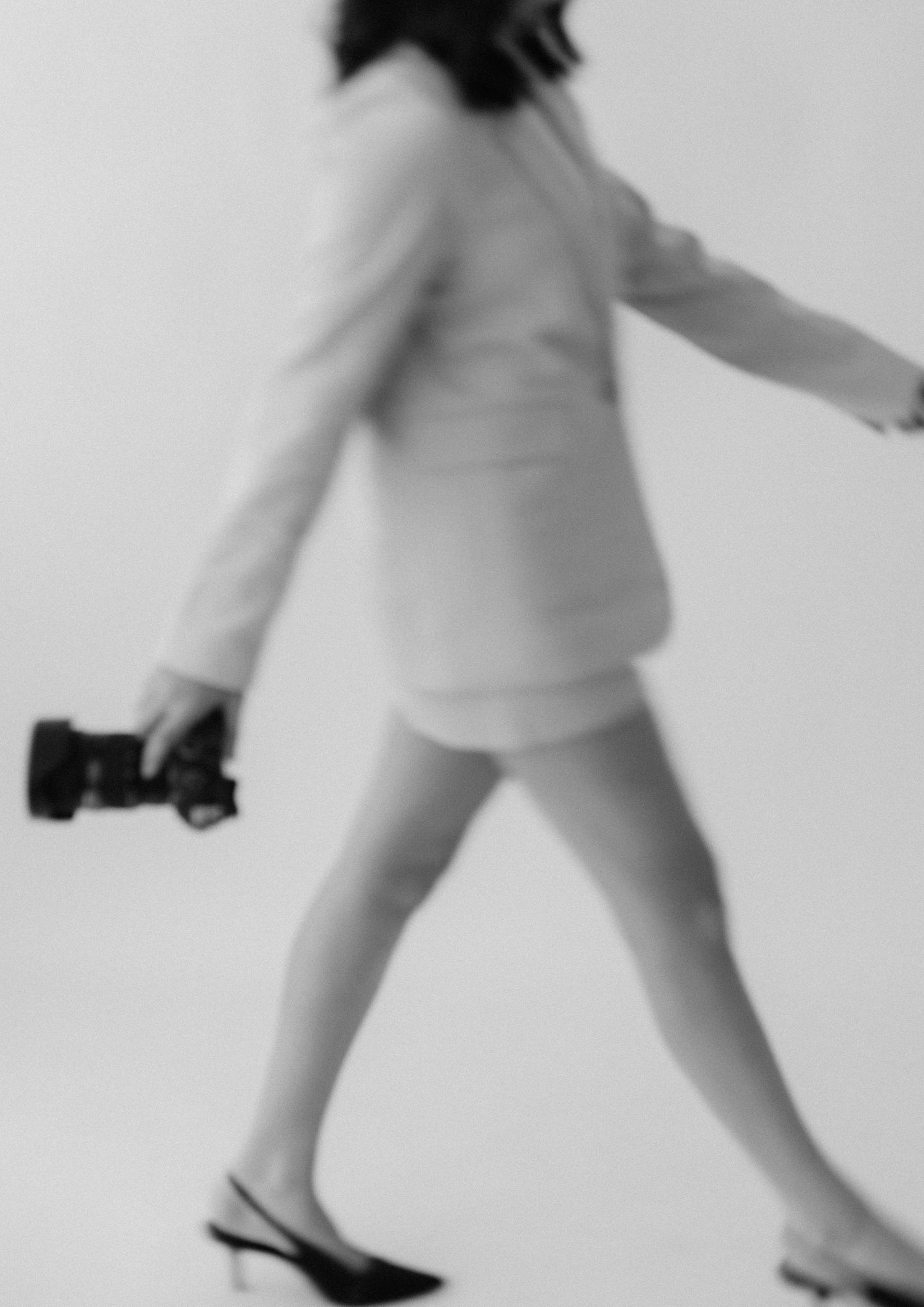 Blurred motion black and white photo of someone walking in a white outfit and slingback shoes while holding a camera.