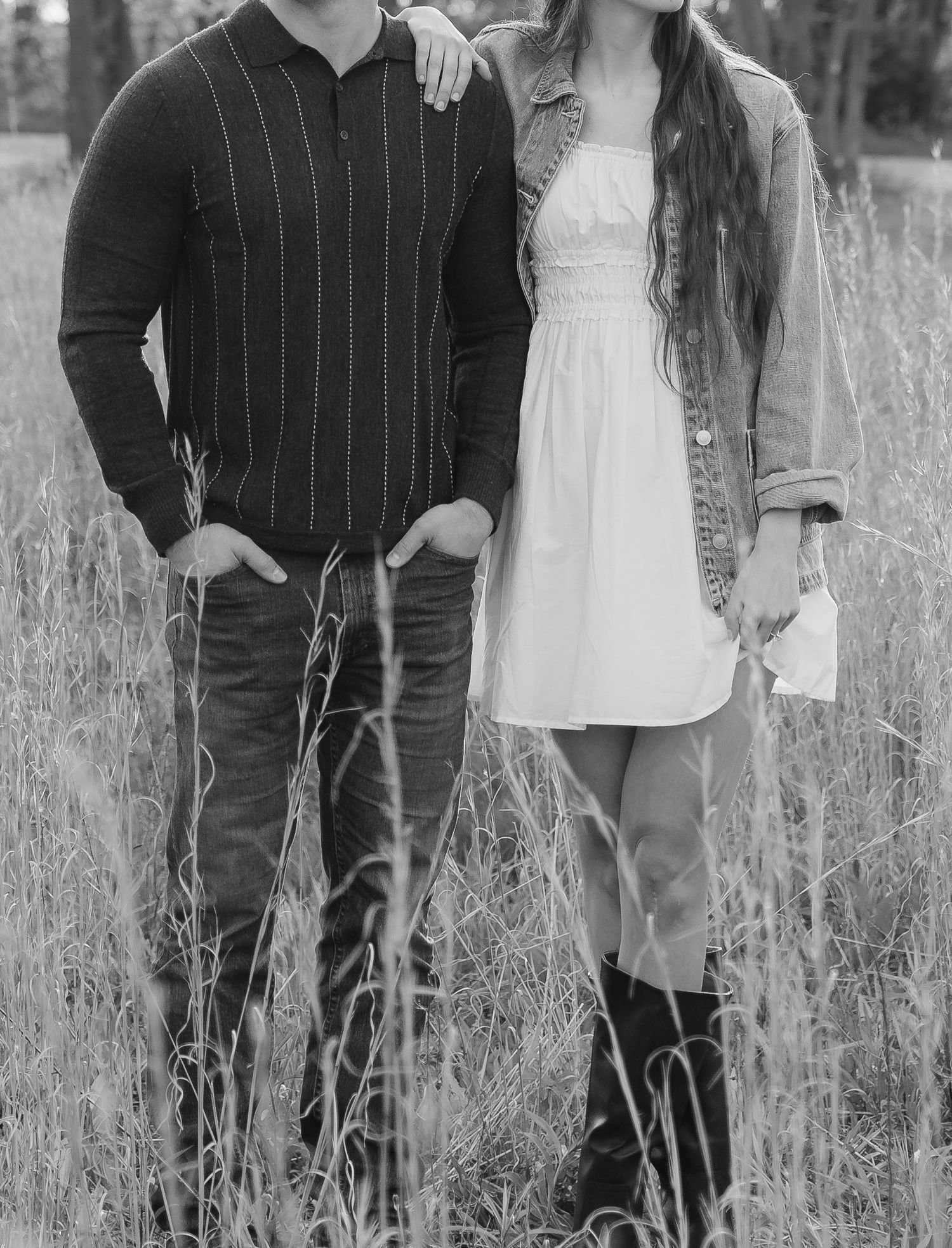 Black and white photo of a couple standing in tall grass, woman in white dress and denim jacket, man in striped sweater.
