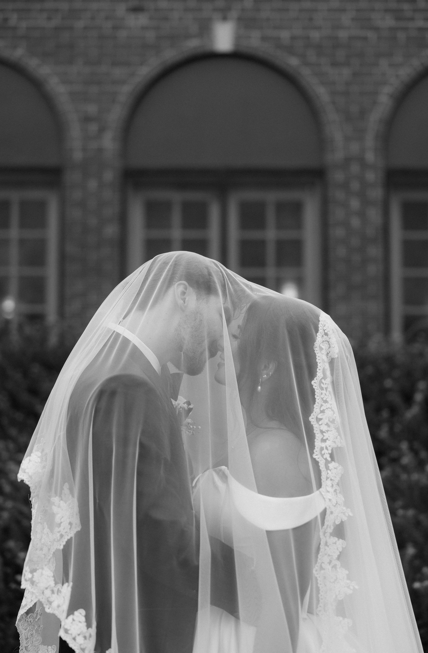 Black and white photo of bride and groom sharing a kiss under a lace-trimmed veil outside a brick building.