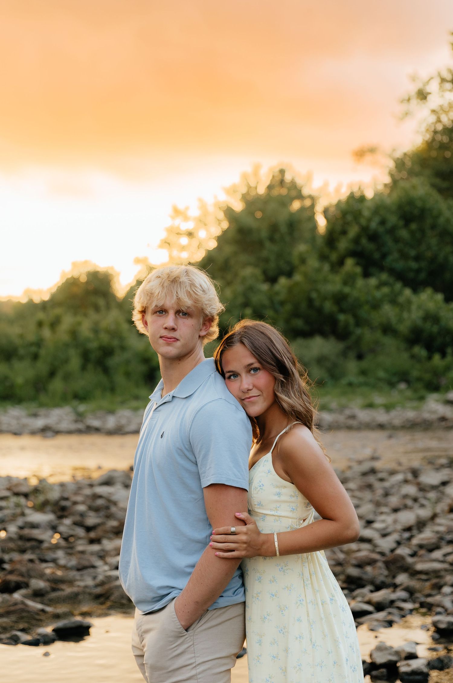 A couple poses against a sunset backdrop near a rocky shoreline.