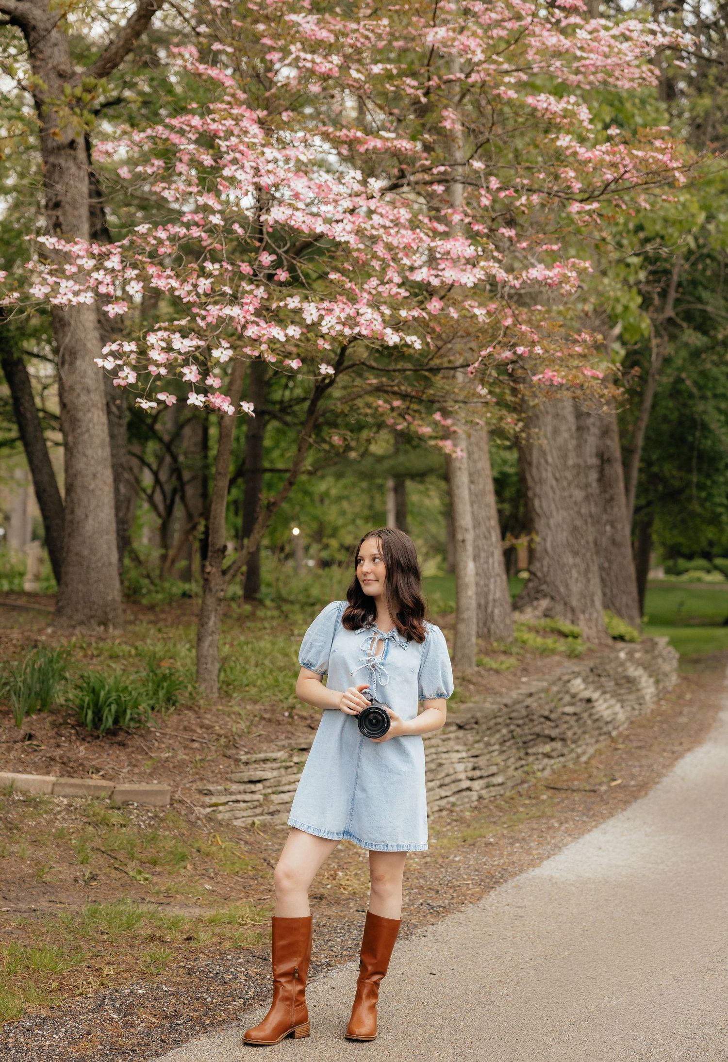Woman in blue dress and brown boots holding camera stands under blooming pink dogwood tree on a garden path.