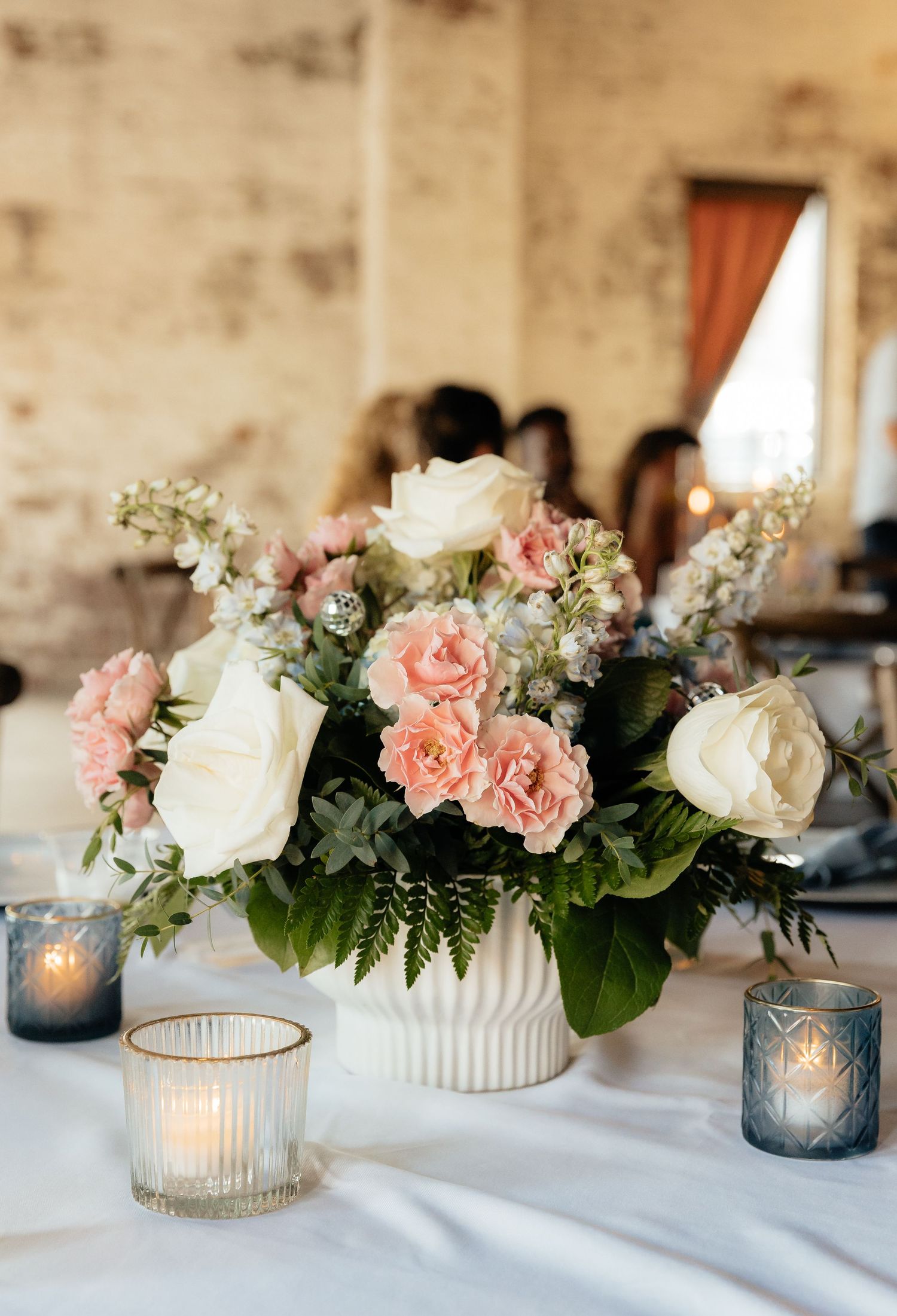 White vase with pink carnations and white roses centerpiece surrounded by blue and clear votive candles on a white tablecloth.