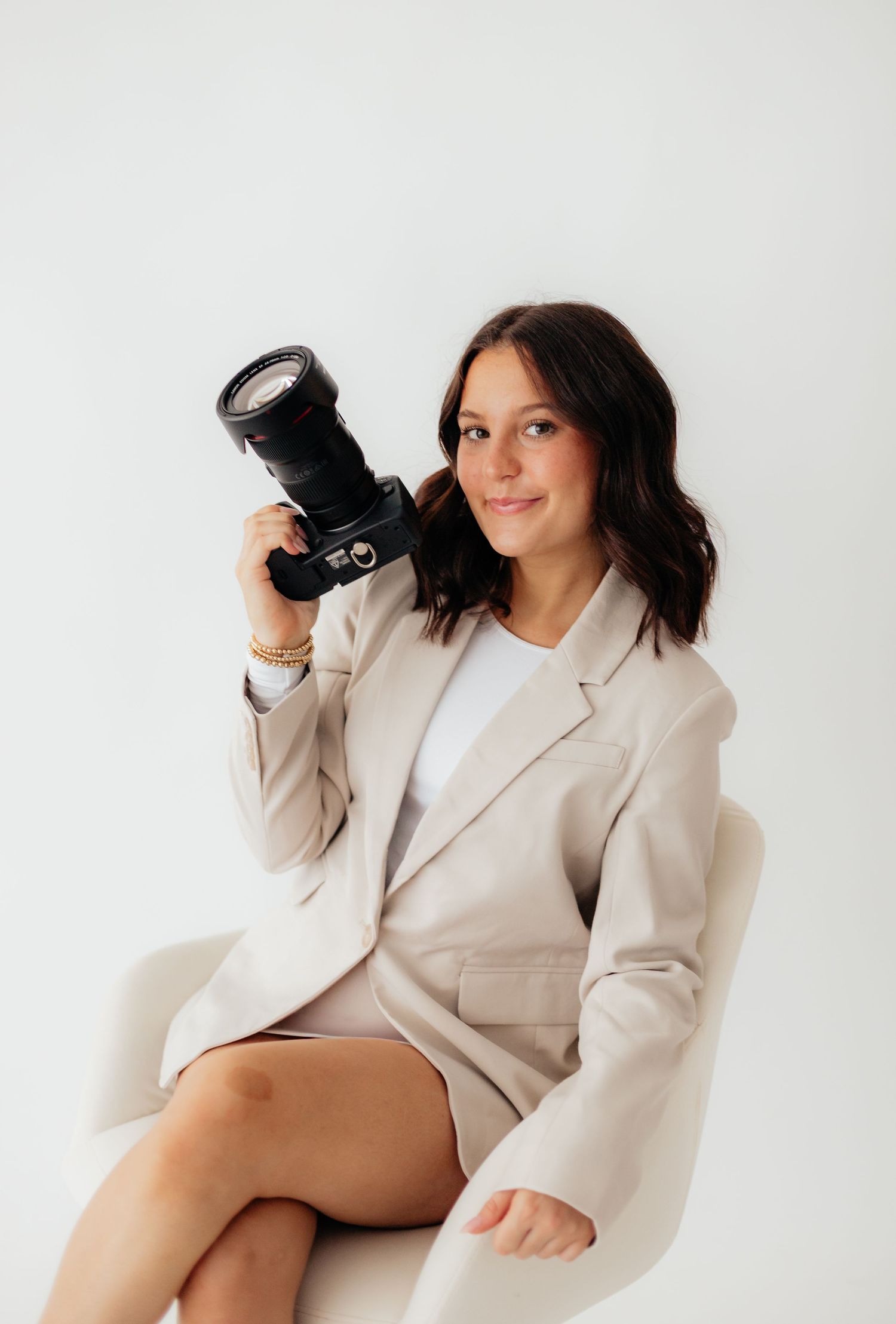 Professional photographer in beige blazer holds DSLR camera while sitting against white backdrop.