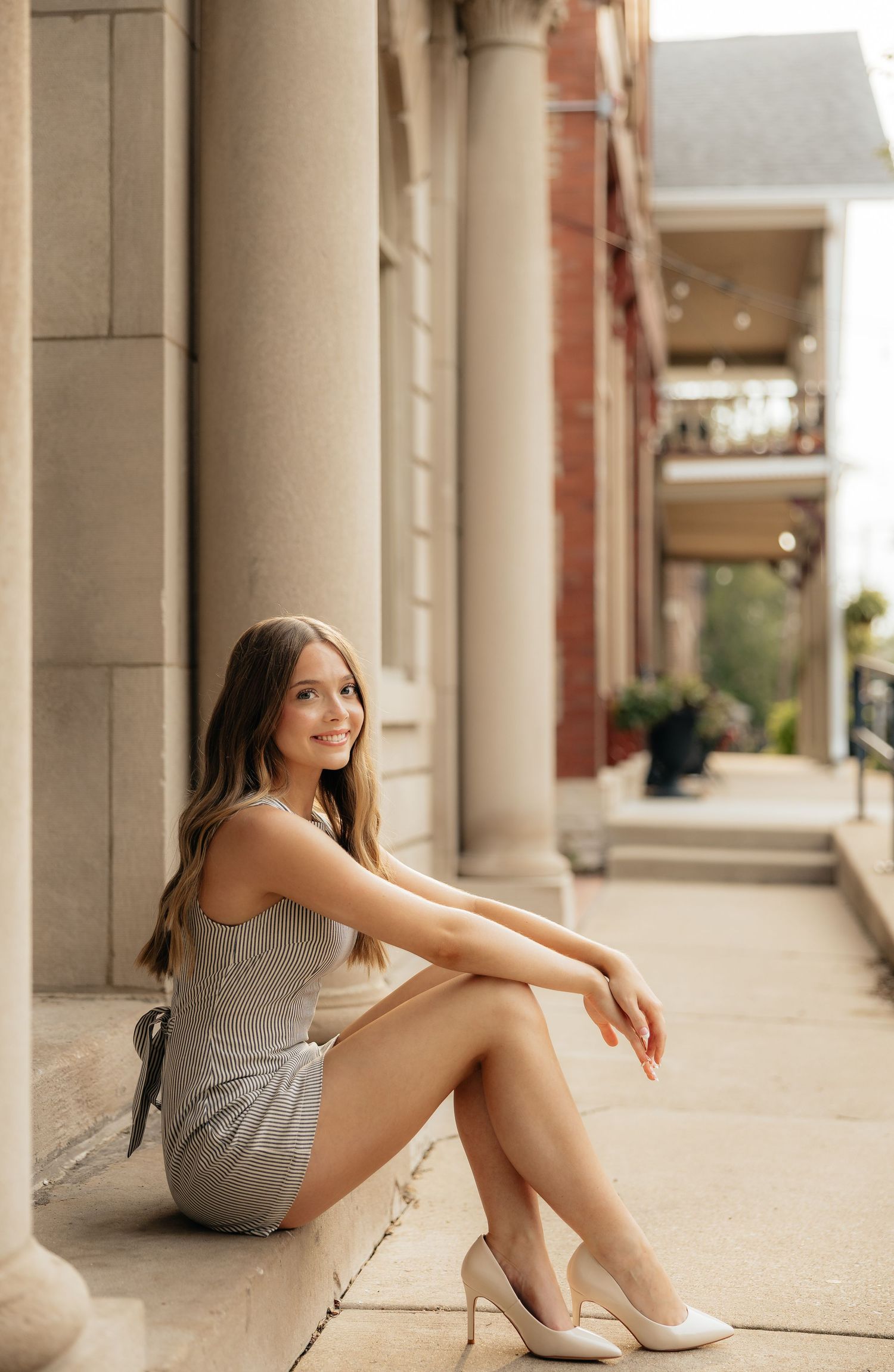 Young woman in gray mini dress and white heels sitting gracefully on stone steps beside classical columns.