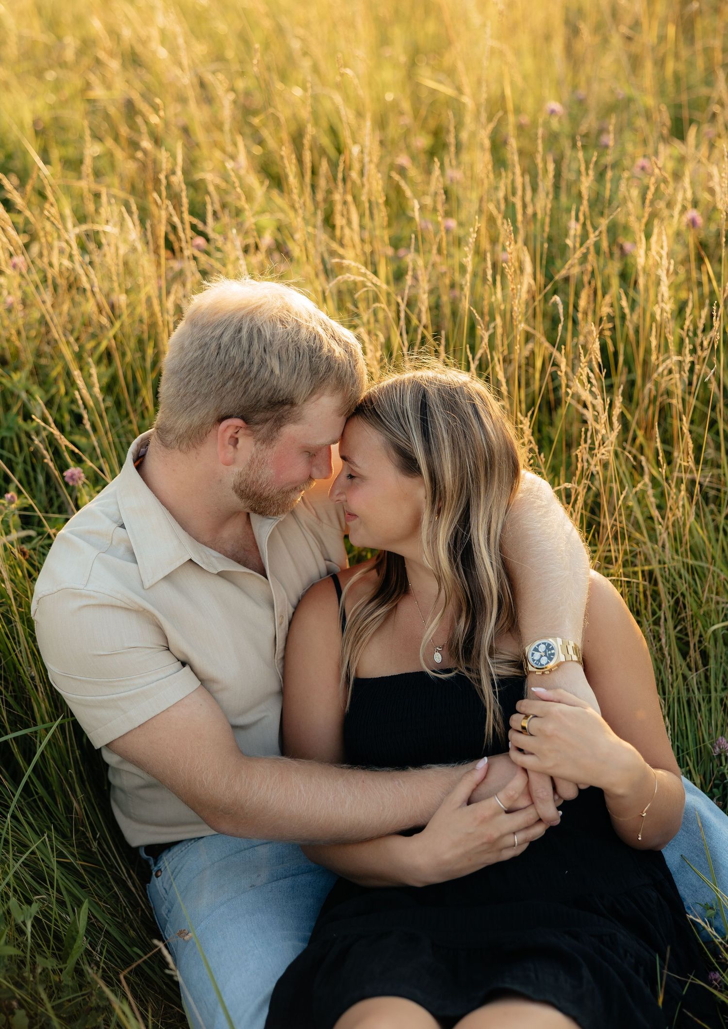 Couple sitting close in tall golden grass field sharing a tender almost-kiss at sunset.