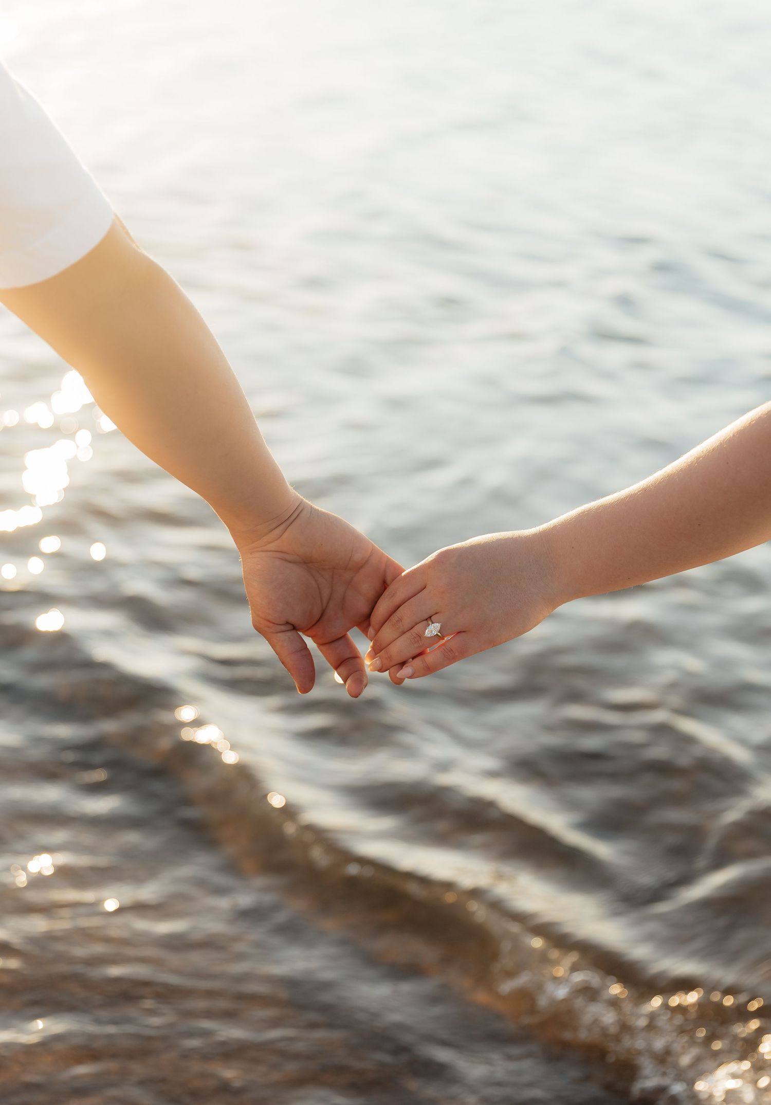 Two people gently holding hands near a sunlit shoreline, one wearing an engagement ring, symbolizing love and romance.