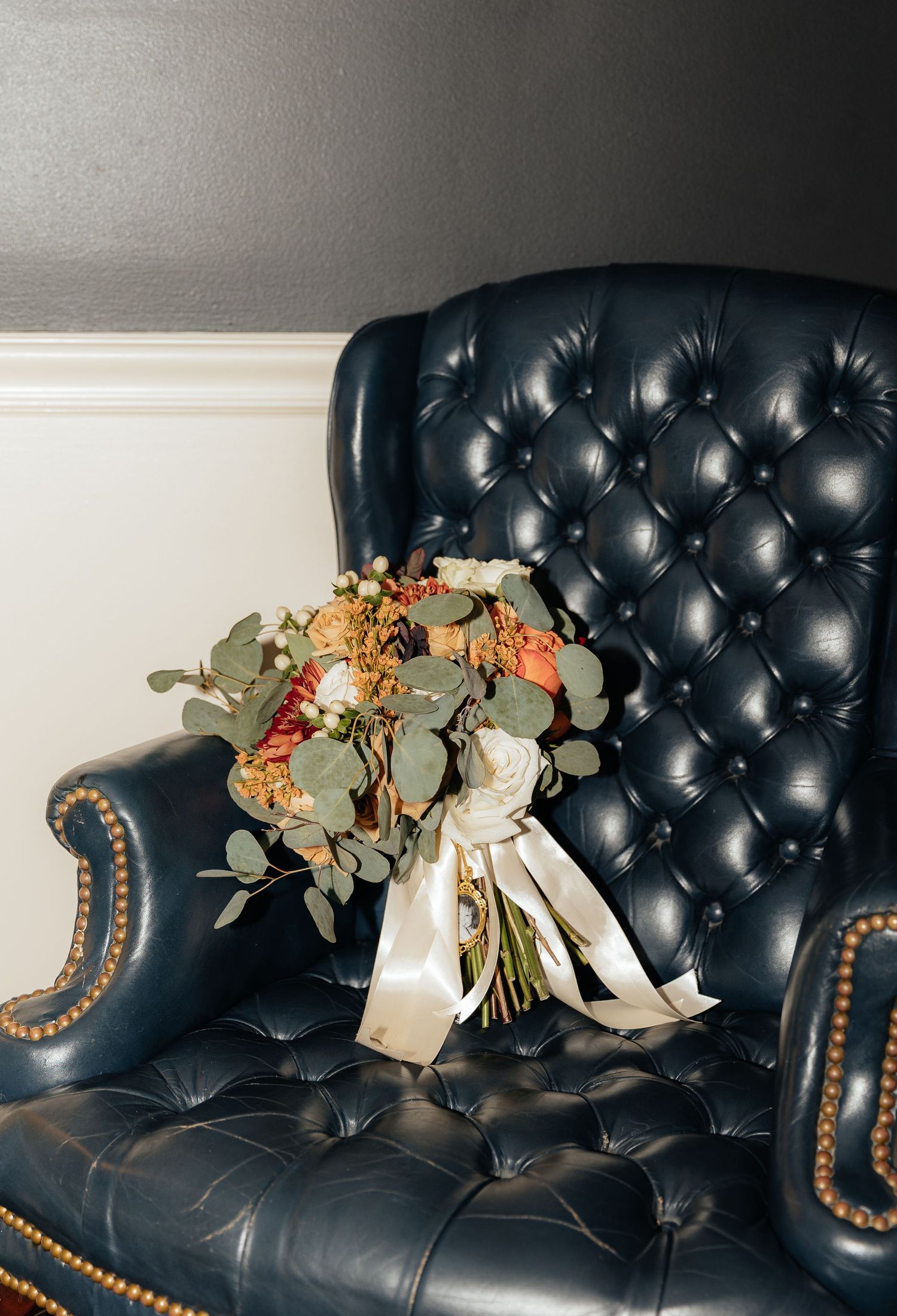 A wedding bouquet with eucalyptus leaves rests on a black leather tufted couch with brass nailhead trim.