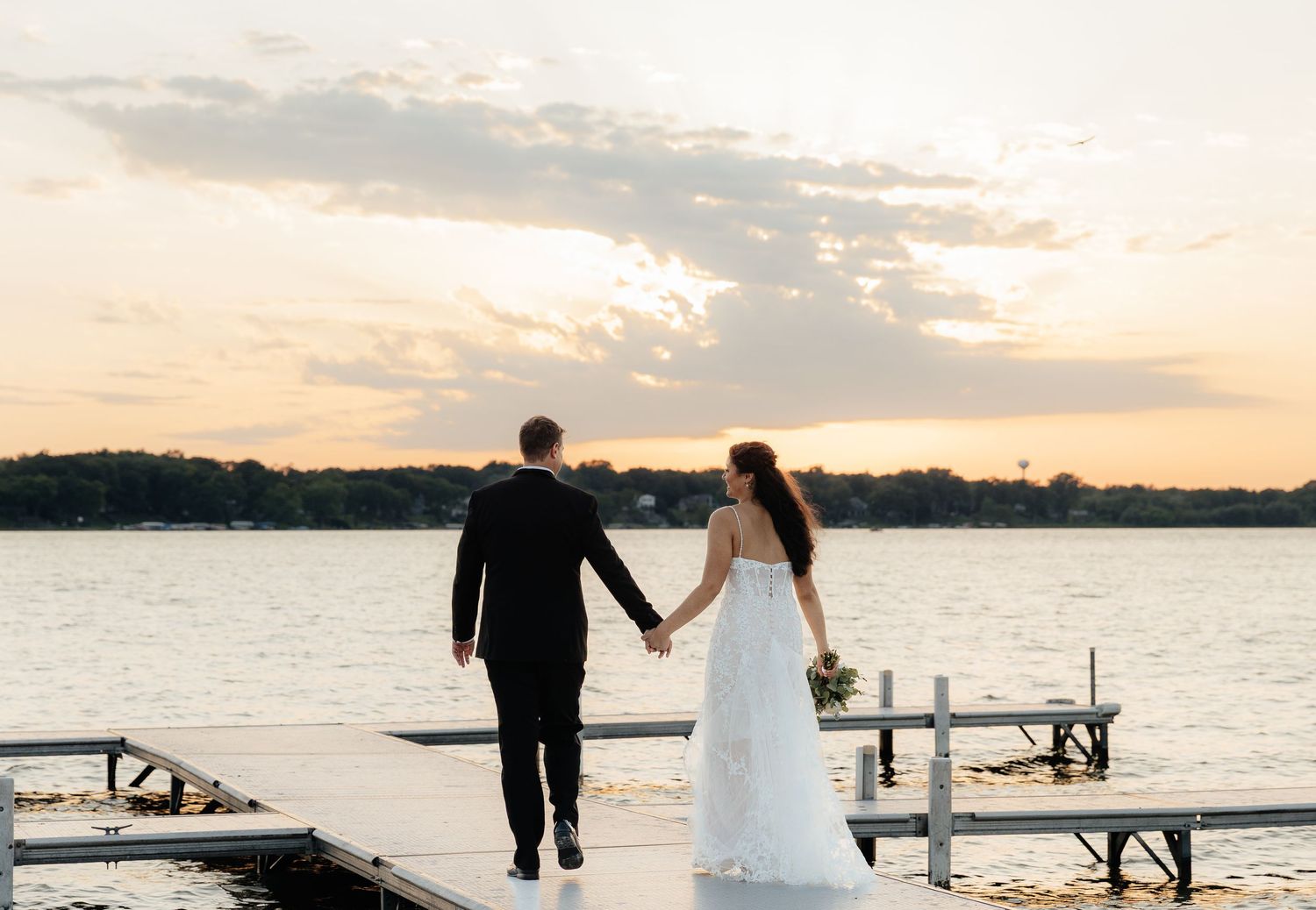 A couple in wedding attire standing on a dock by the water during sunset creating a romantic silhouette.