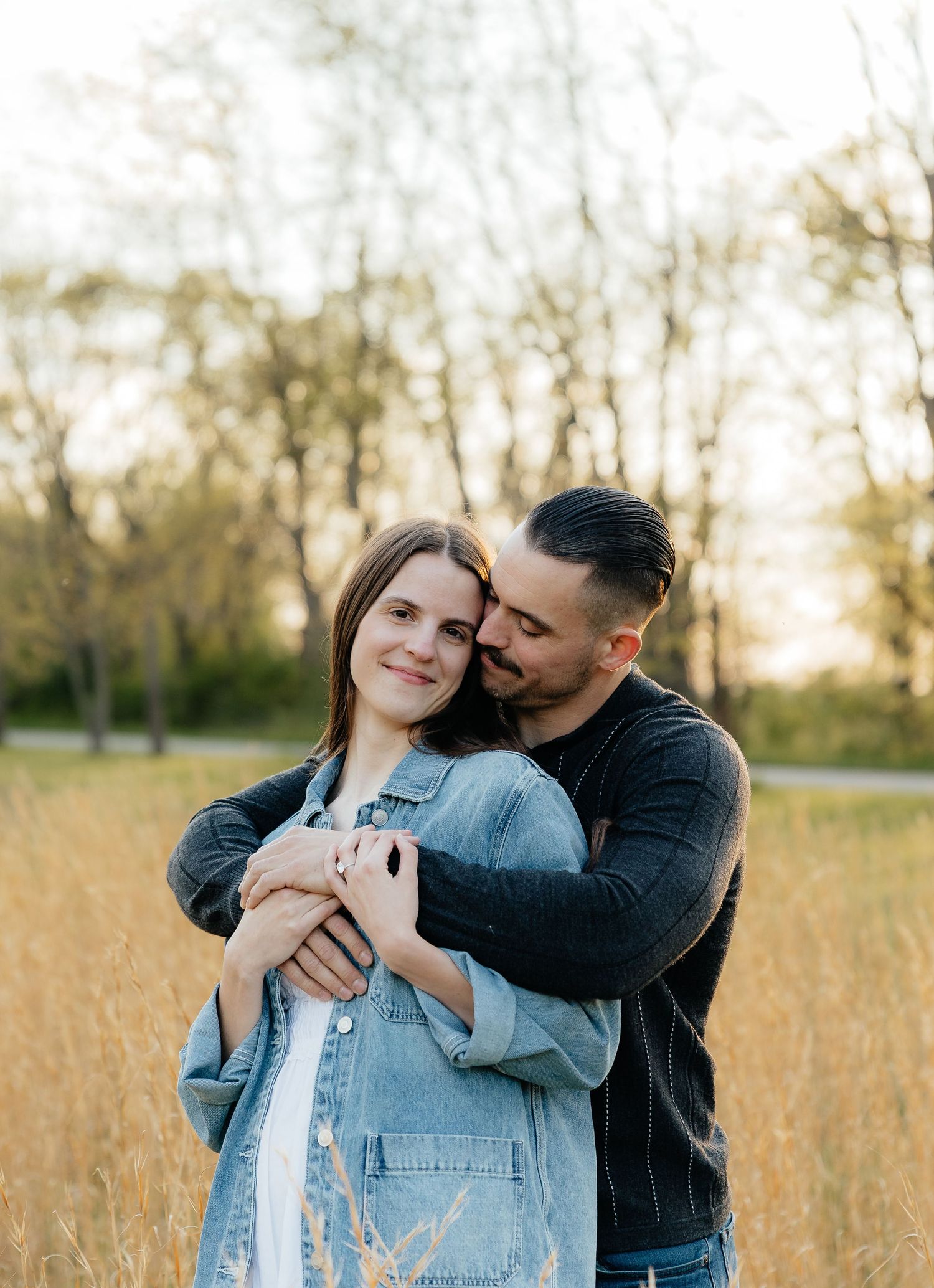 A couple embraces in a golden field at sunset, woman in denim jacket smiling as man holds her from behind.
