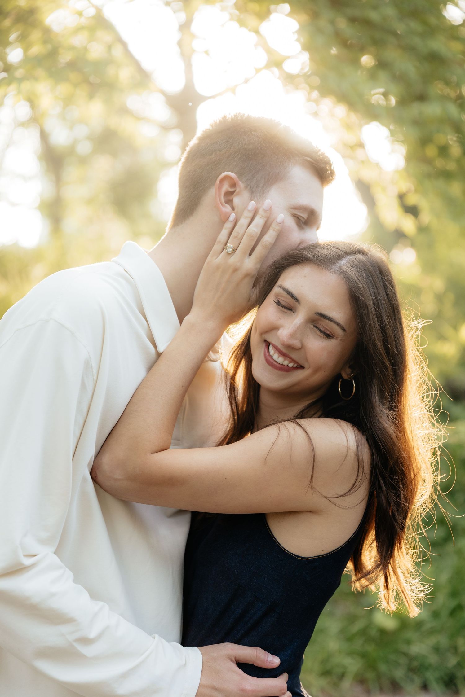 Happy couple embracing outdoors at sunset, woman smiling with eyes closed, man hugging her from behind in golden light.