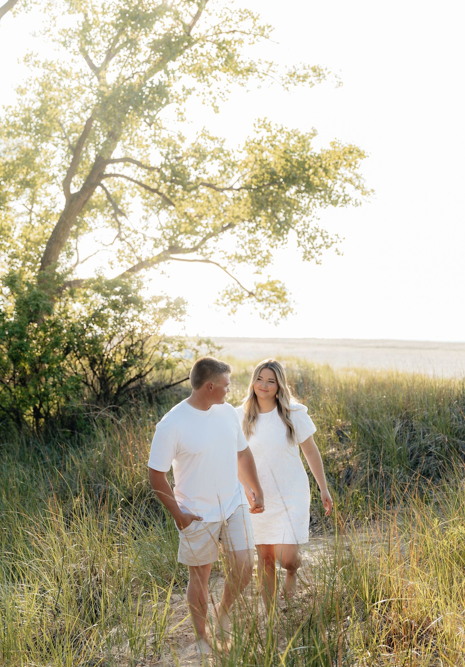 Couple in white outfits holding hands walking through golden marsh grass at sunset near the beach.
