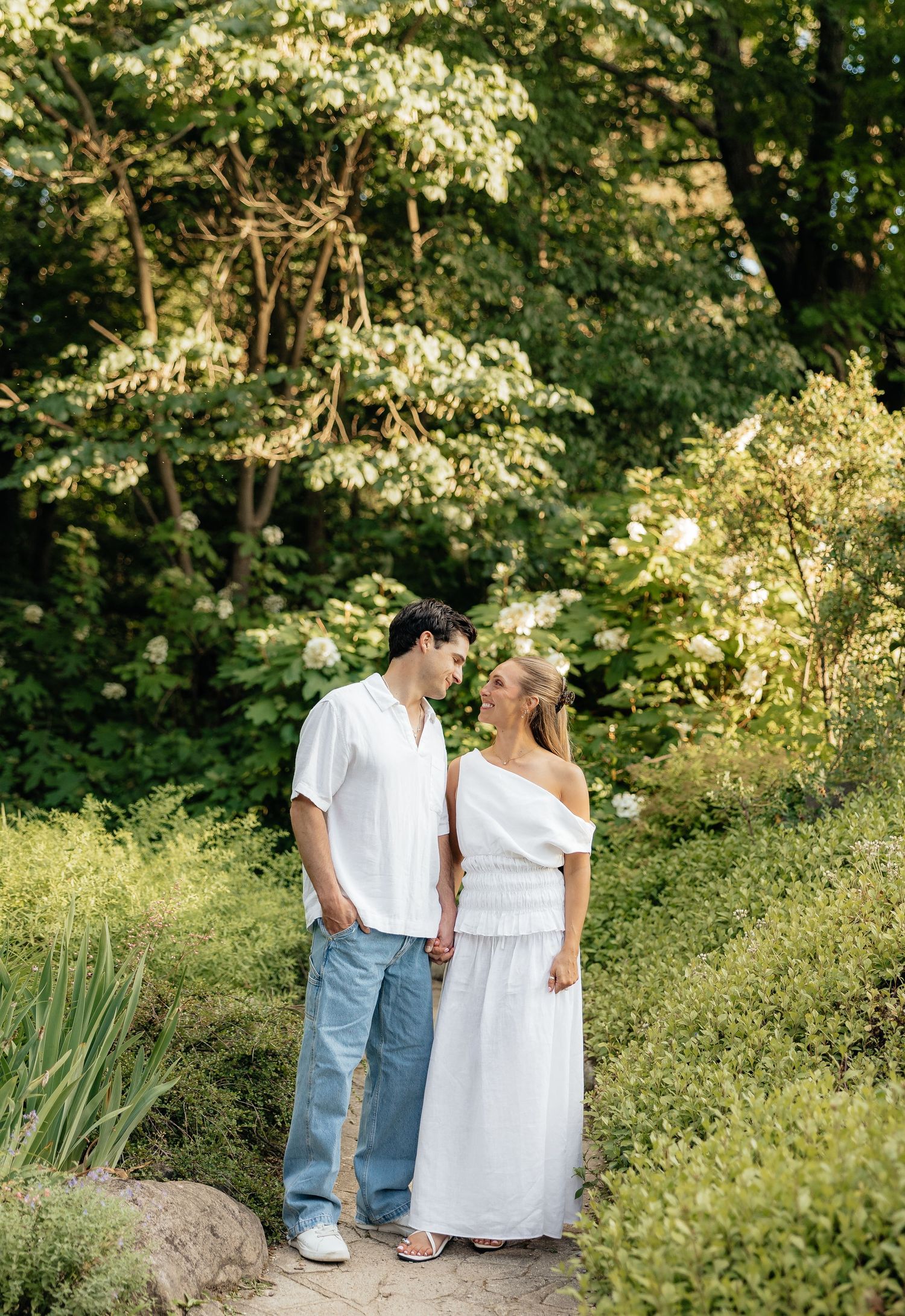 Couple in white outfits holding hands on a garden path surrounded by lush green trees and white flowering shrubs.