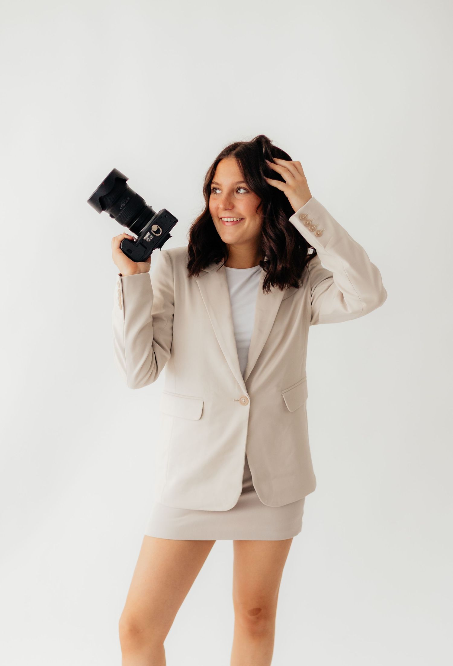 A photographer in a beige blazer and white sweater poses playfully with a camera in a minimalist studio setting.