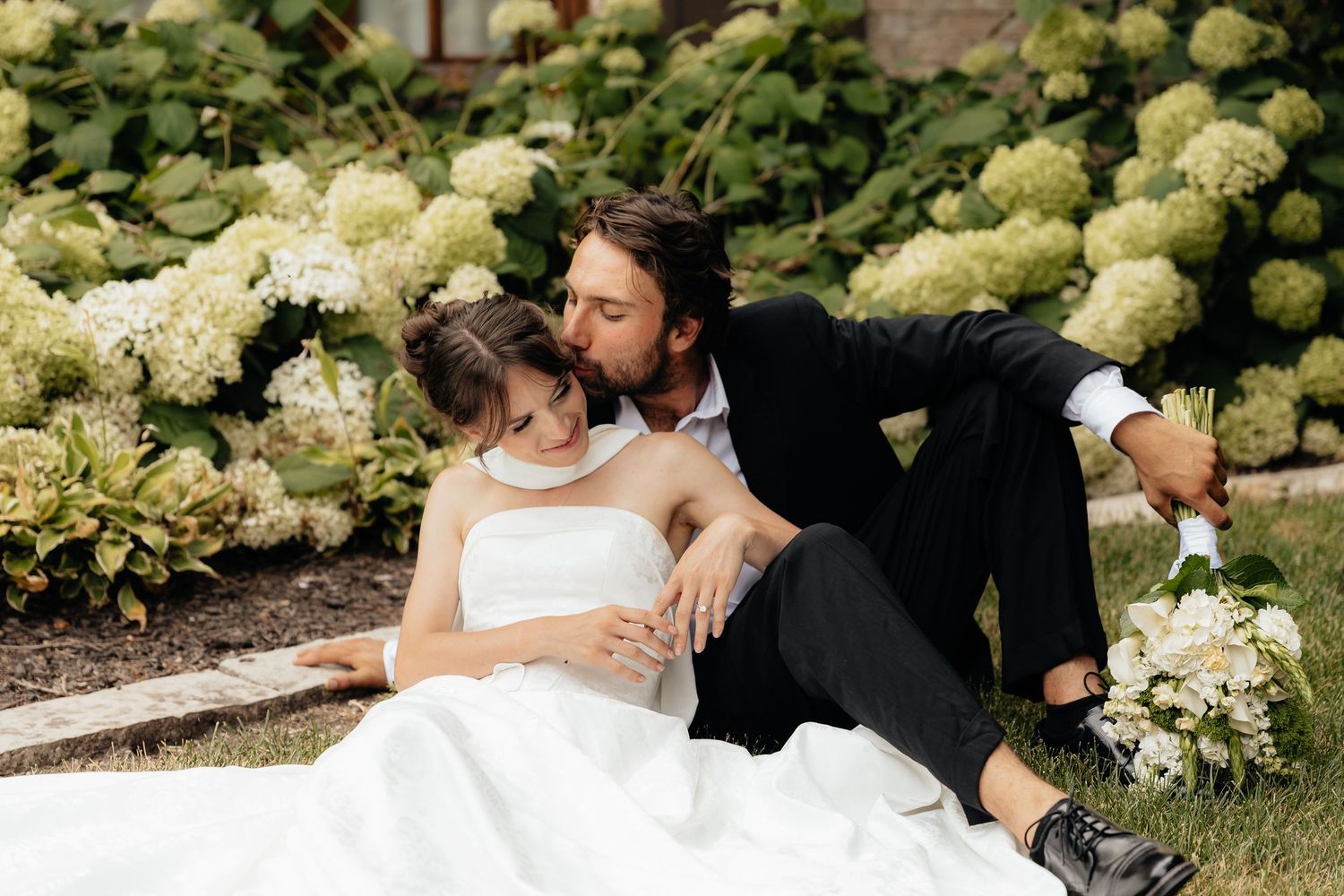 A romantic wedding couple embraces by a garden of white hydrangea bushes on their special day.