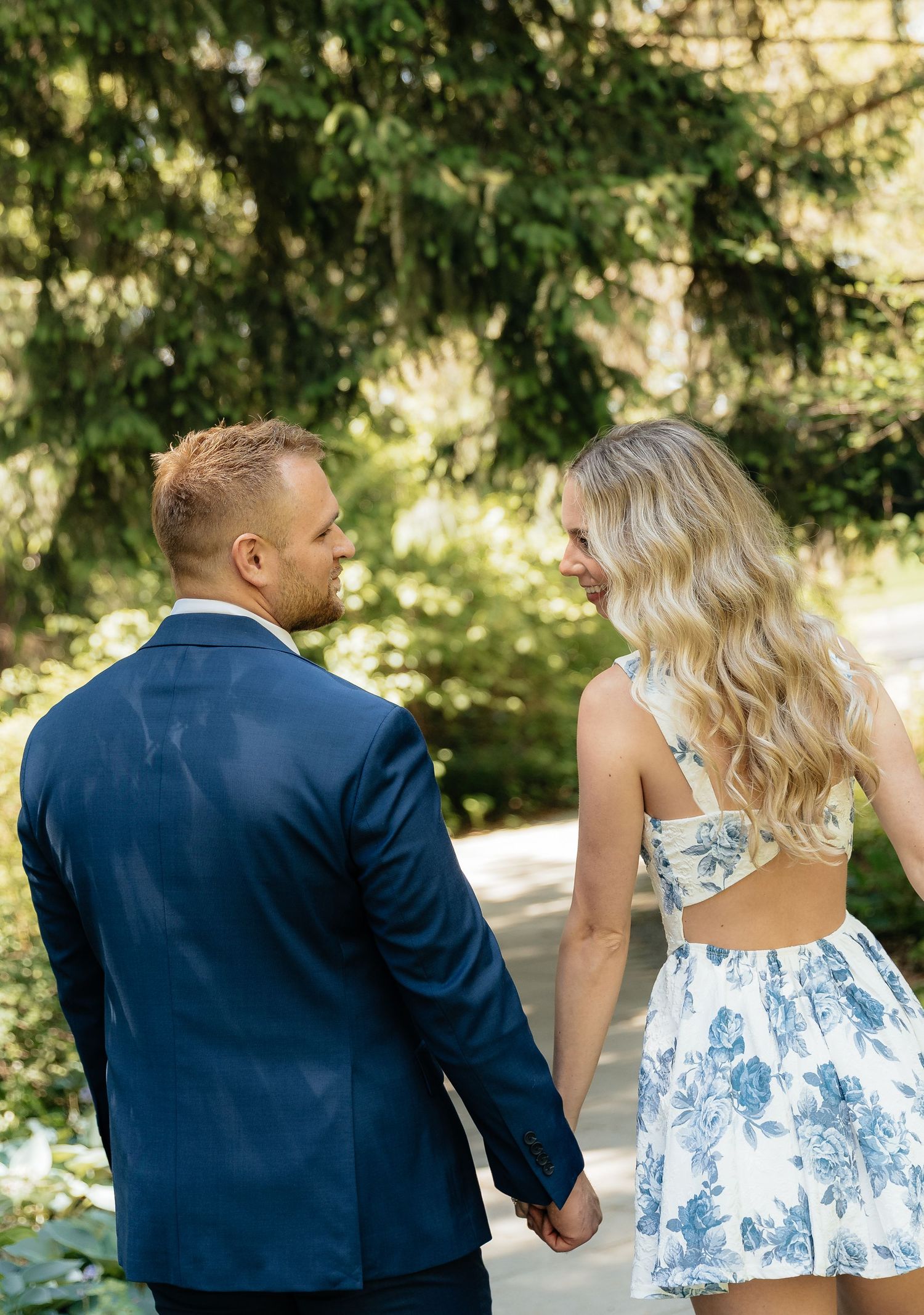 A couple holding hands walks through a lush garden, man in navy suit, woman in floral sundress with cutout back.