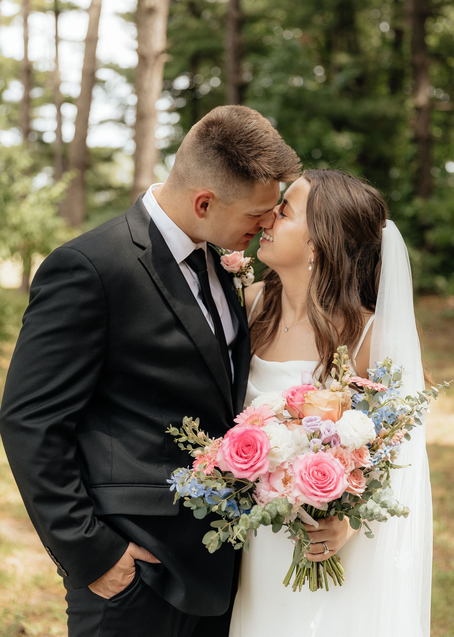 Bride and groom touching foreheads, sharing an intimate moment outdoors, bride holding a pink and blue floral bouquet.