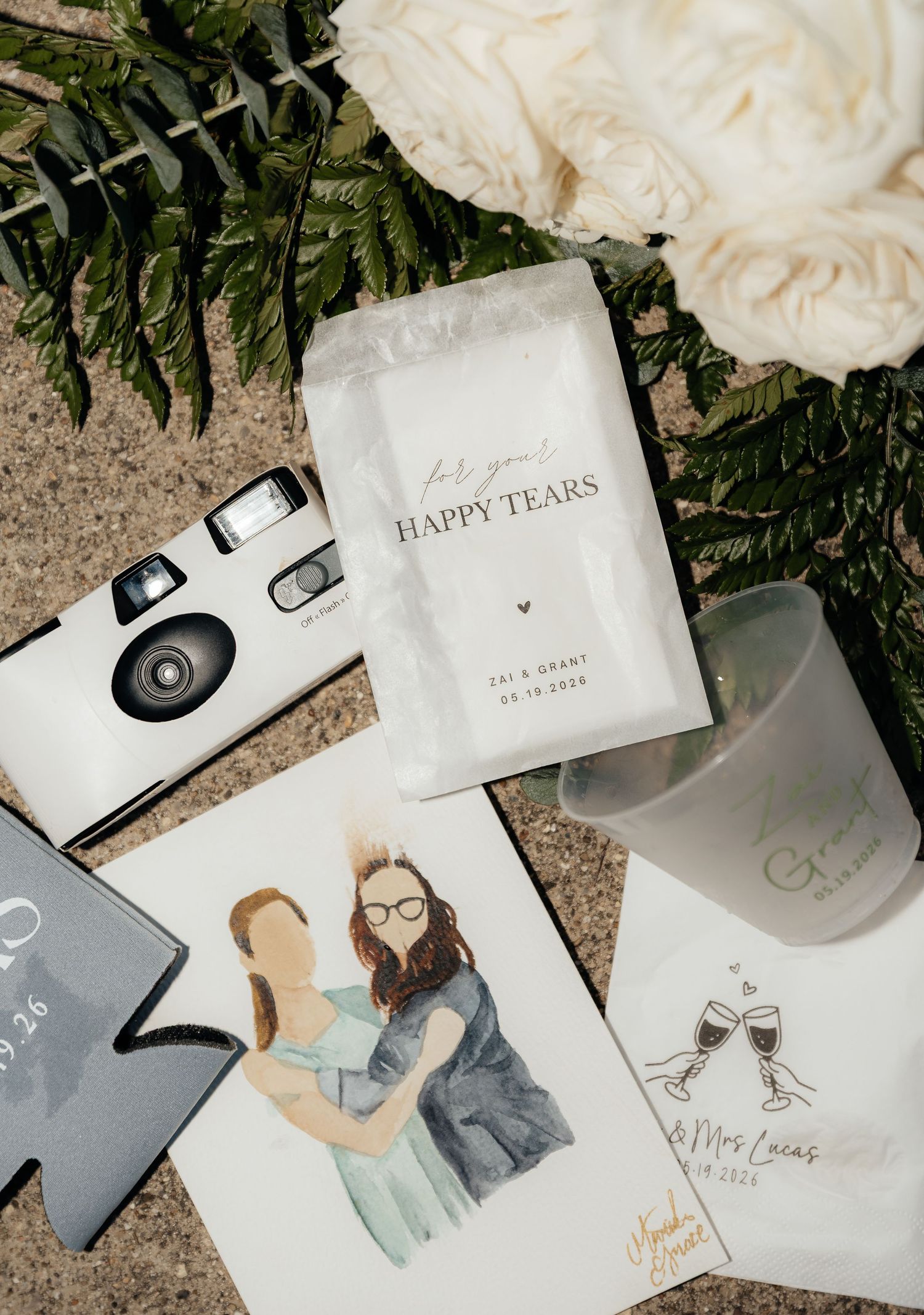 Wedding flat lay with disposable camera, happy tears packet, couple portrait, and personalized napkins on neutral background.