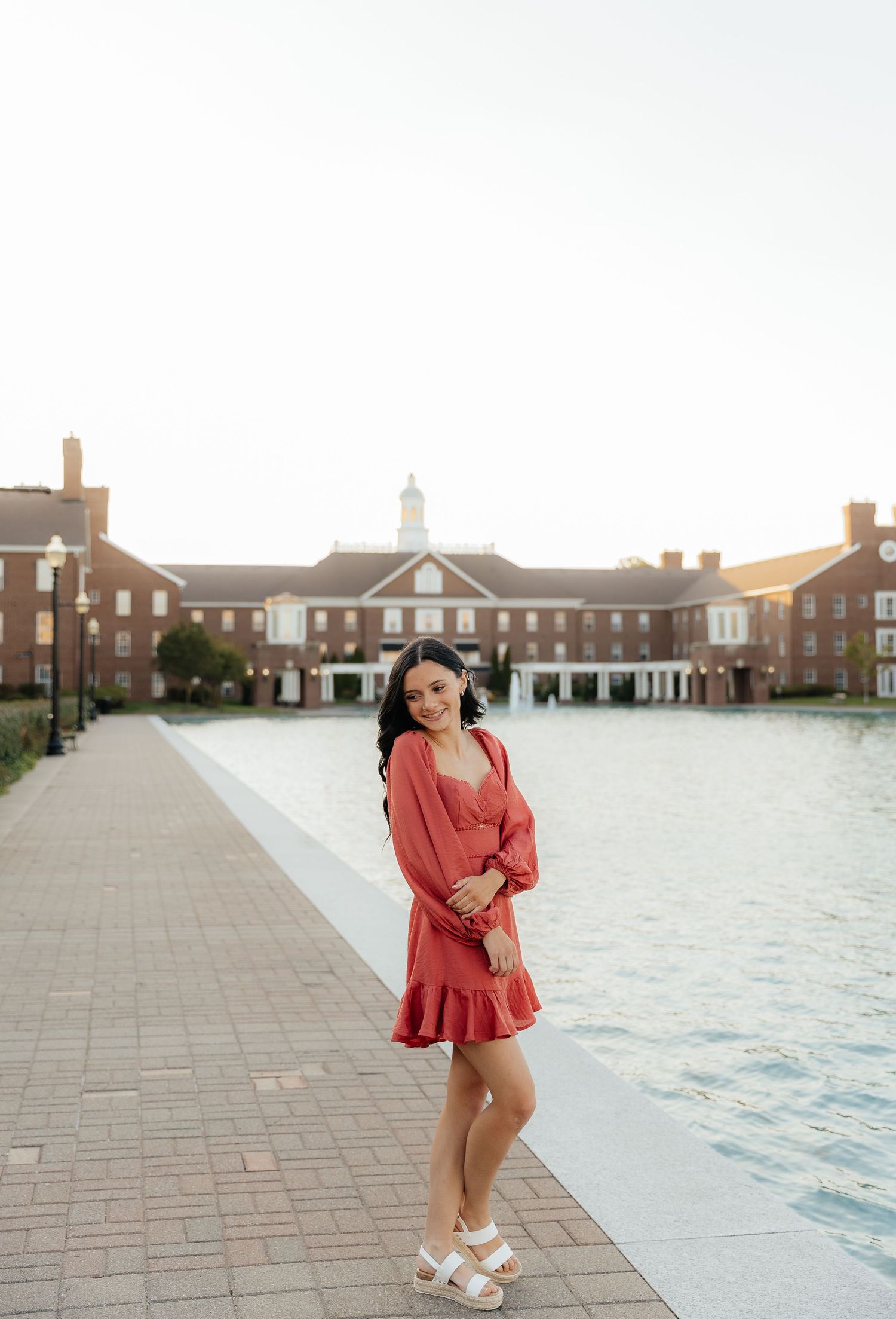 A person in a coral-colored romper poses playfully in front of a brick colonial-style building on a sunny day.