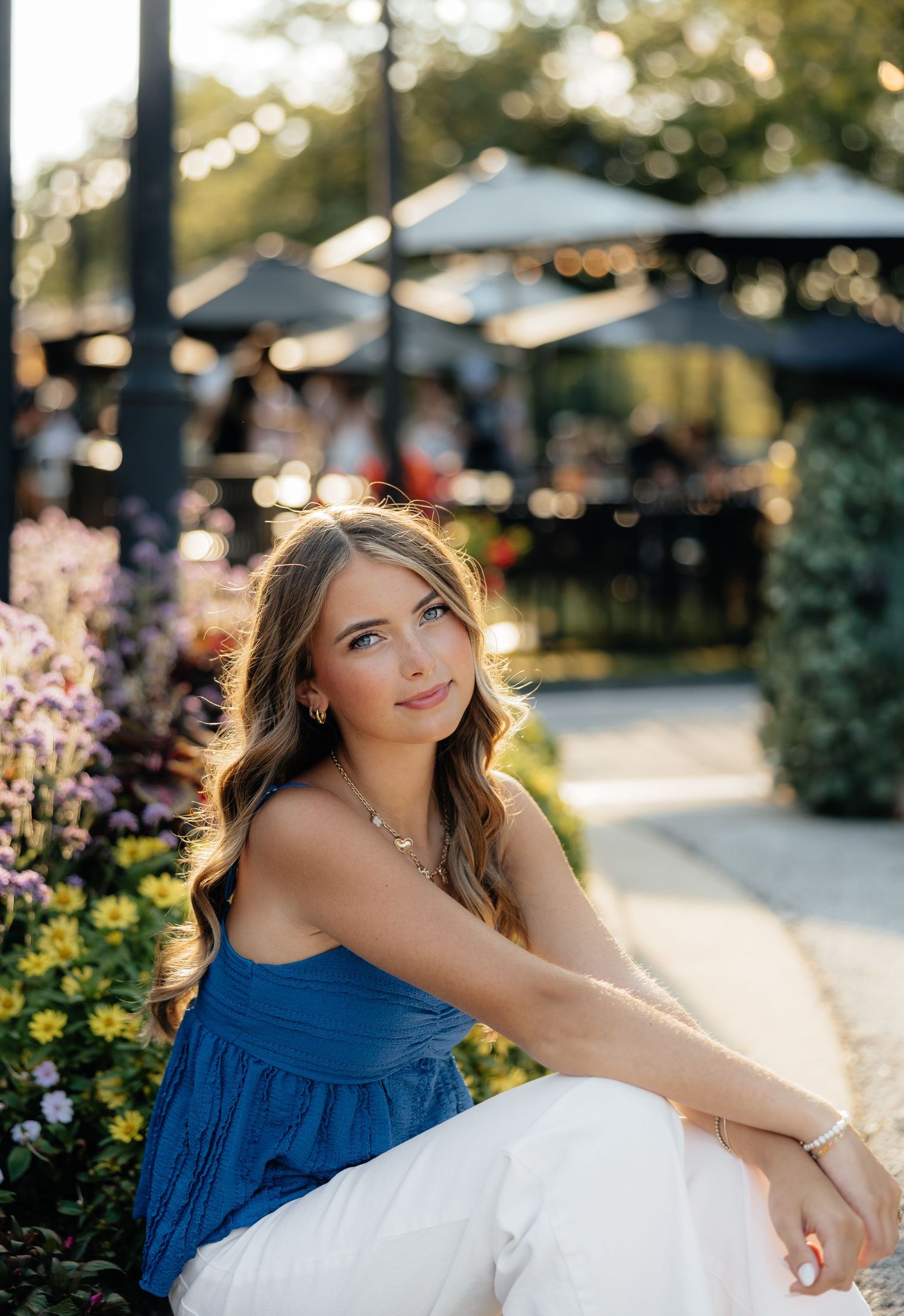 Young woman in blue top and white skirt sitting by colorful garden flowers in warm golden hour light.