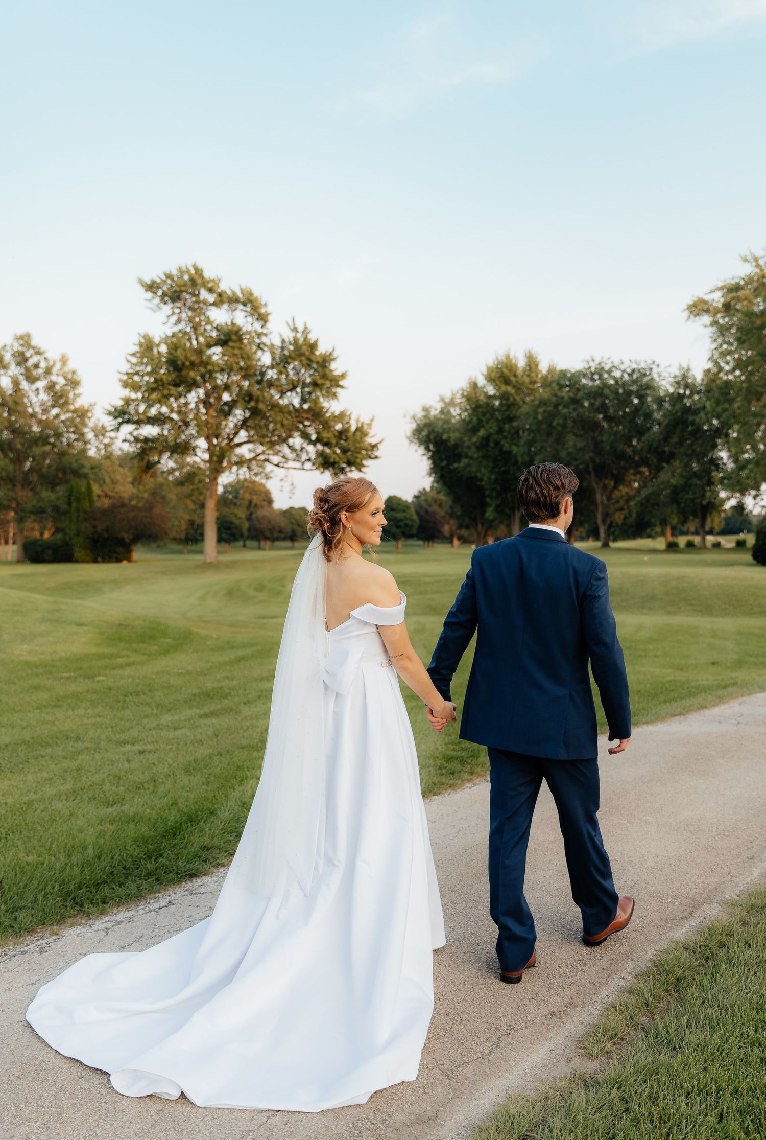 Couple walks along path on golf course during wedding, bride in white gown and groom in navy suit holding hands.