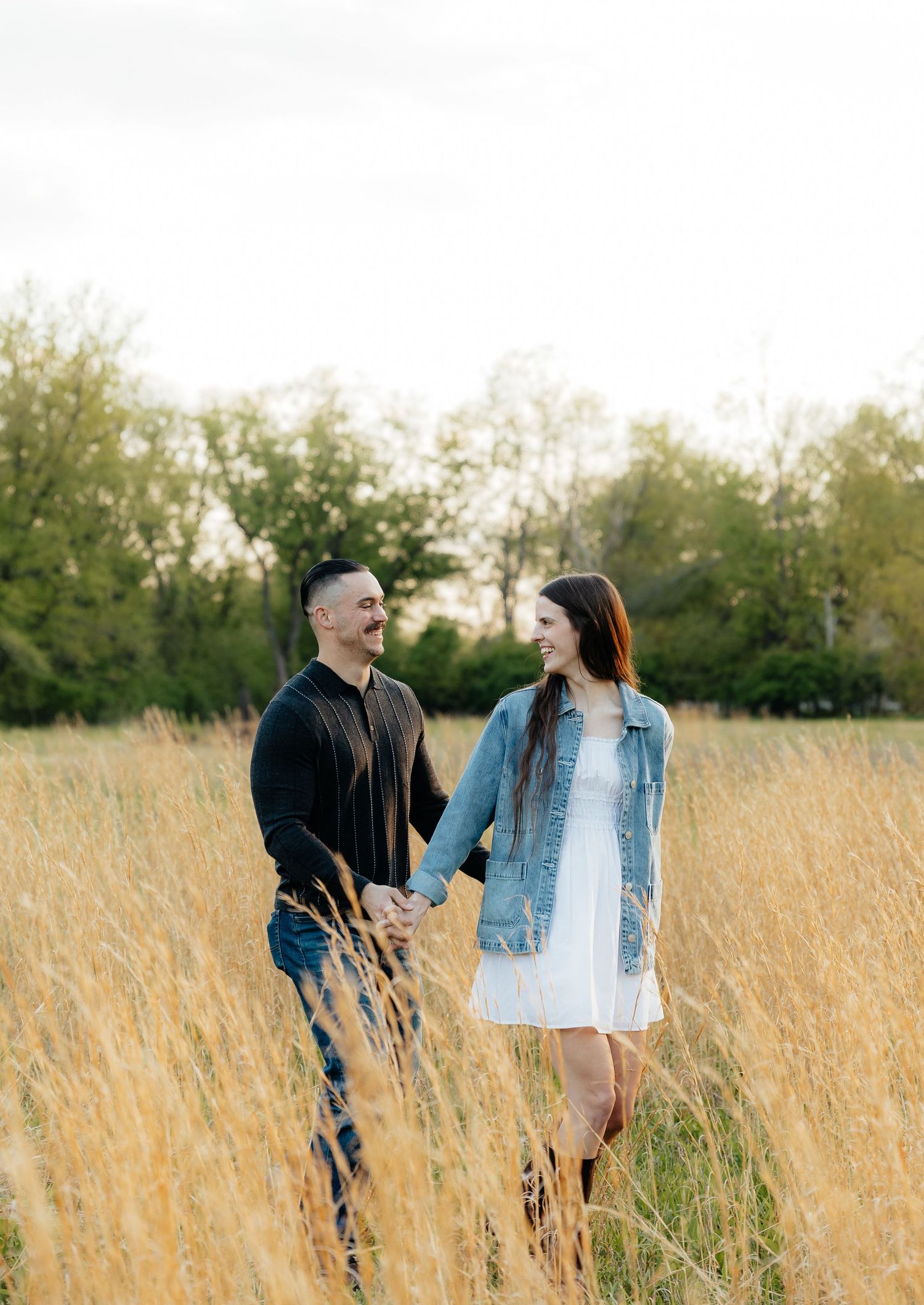 Couple holding hands and smiling in a golden field, woman in white dress and denim jacket, man in black outfit.