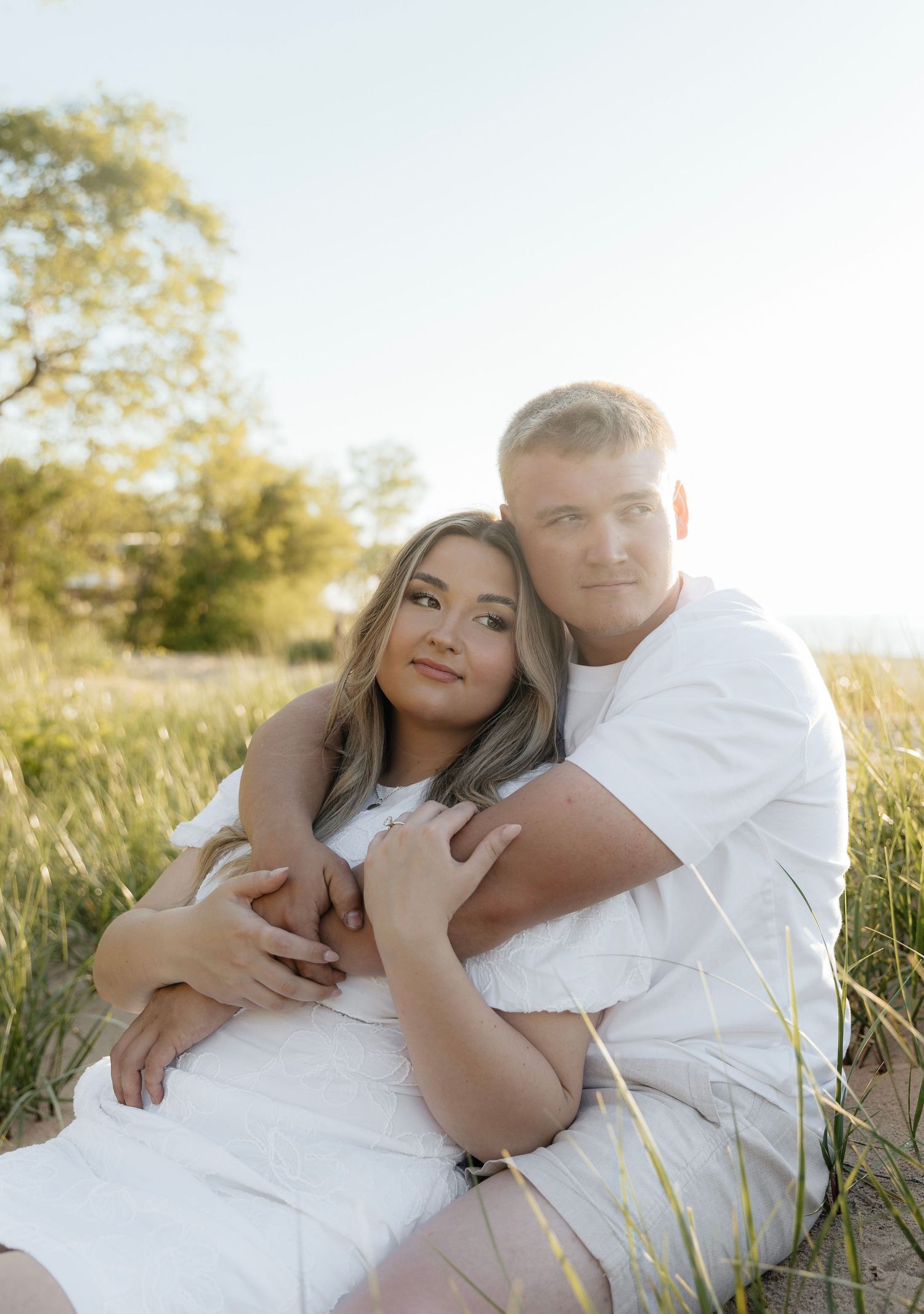 Couple in white outfits sitting in a sunny field, woman leaning back into man's arms, golden hour light.