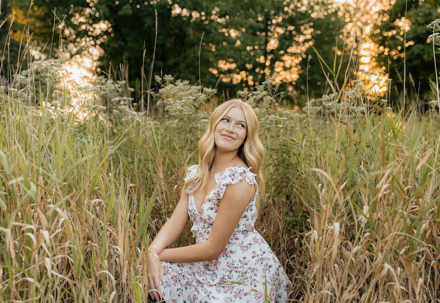 Woman in floral dress poses in golden field of tall grass during sunset.