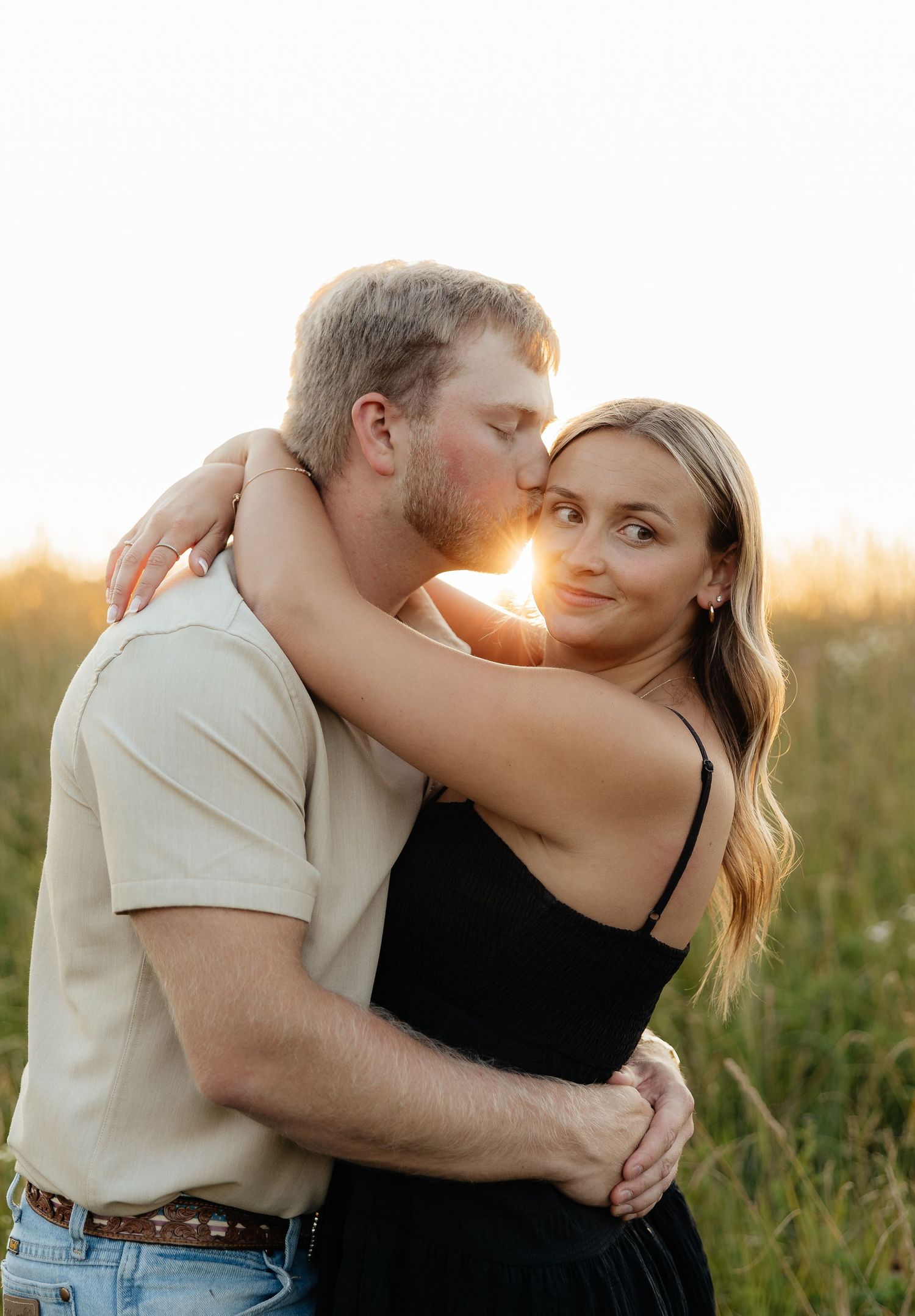 Couple embracing at golden hour in a field, man kissing woman's cheek as she smiles softly at camera.