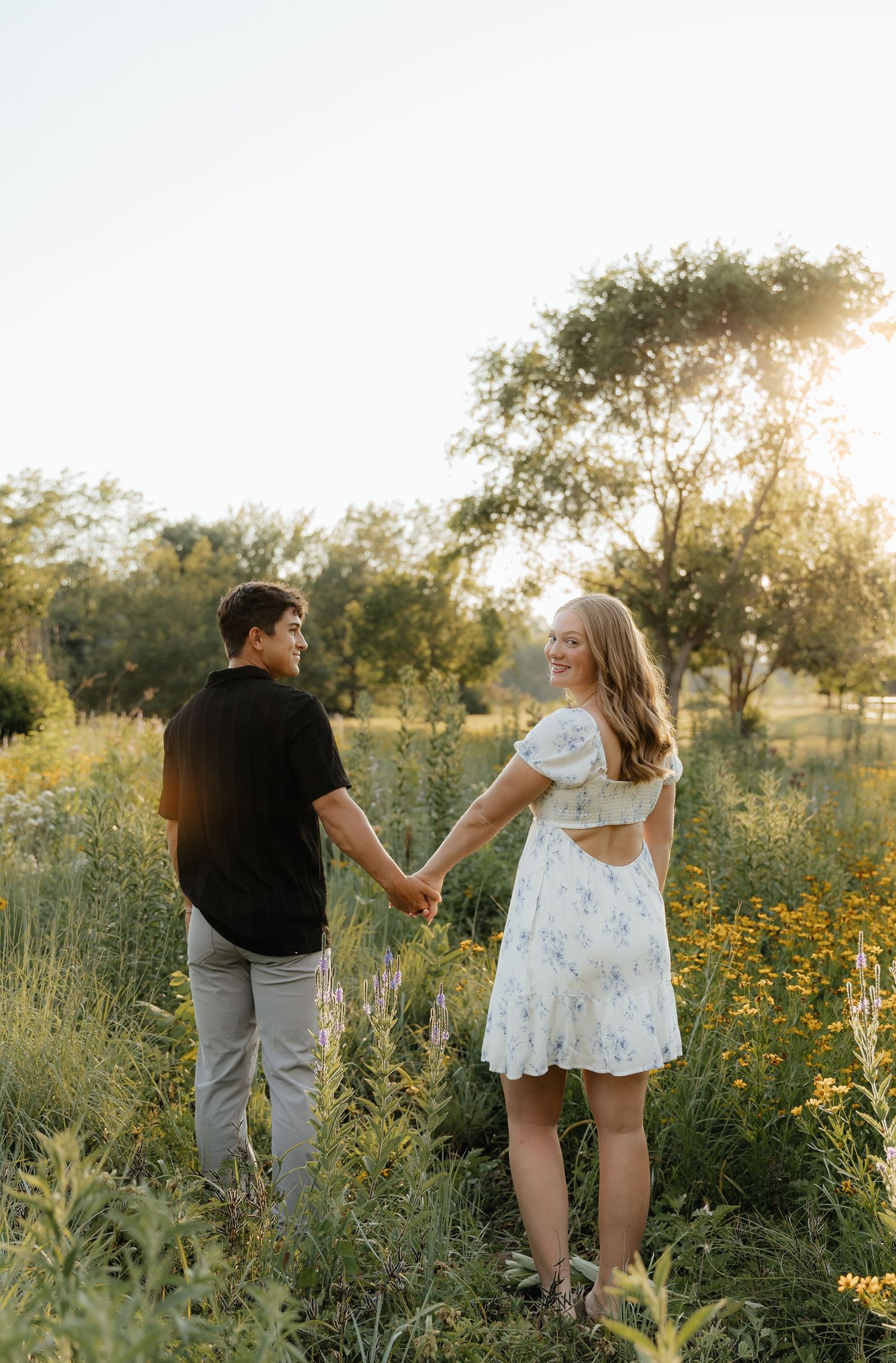 A couple holds hands while walking through a wildflower field at sunset.