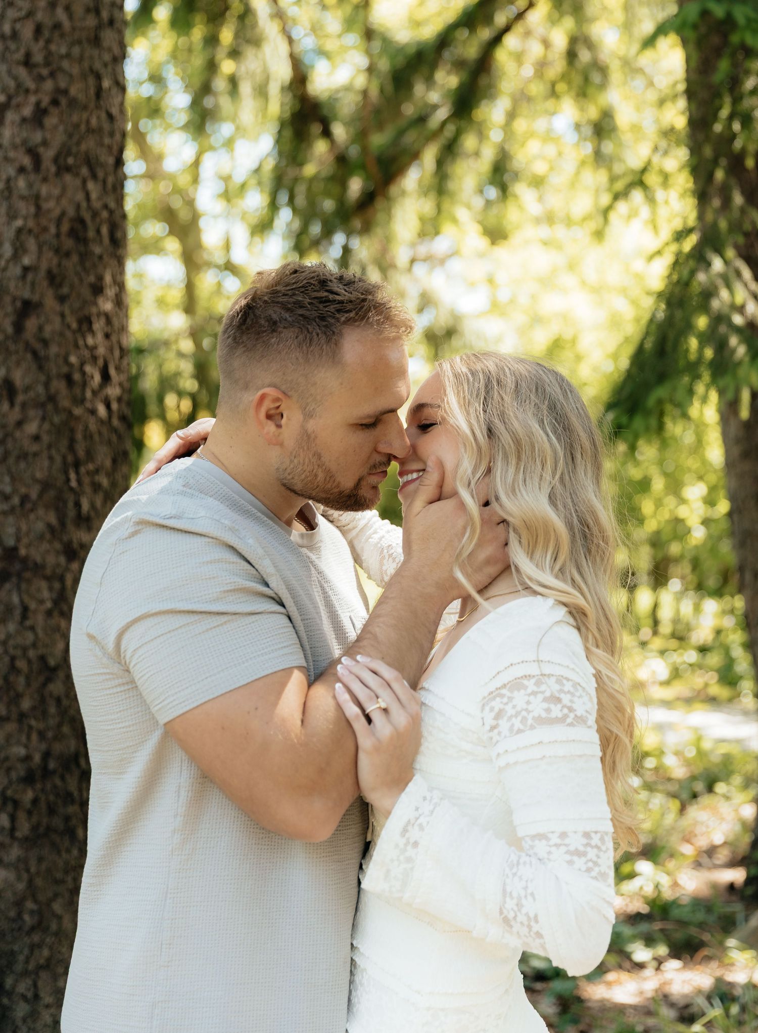 Couple sharing a romantic almost-kiss outdoors among tall green trees, man gently holding womans face.