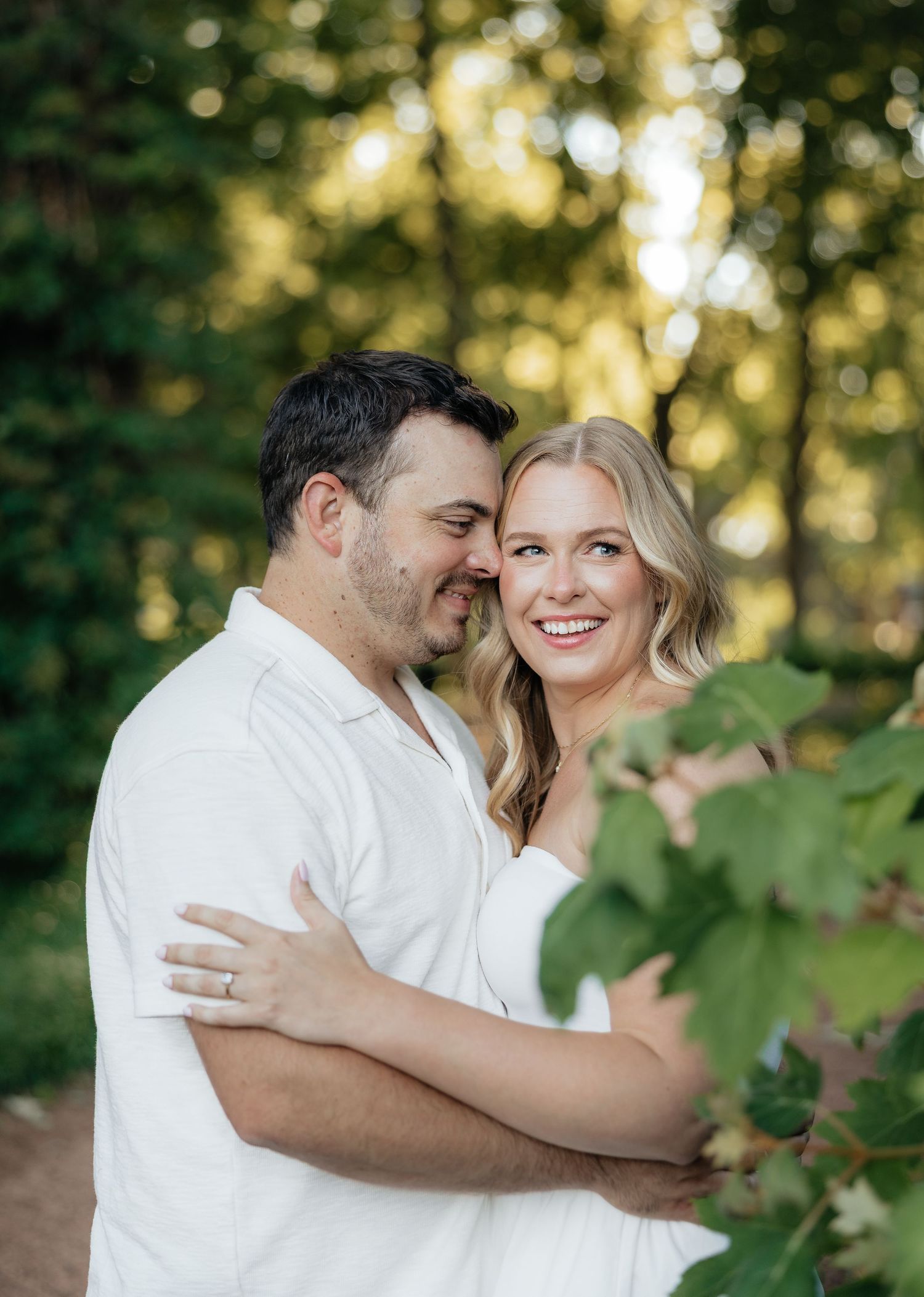 Couple in white outfits embracing outdoors among green trees, woman smiling warmly, engagement ring visible on her hand.