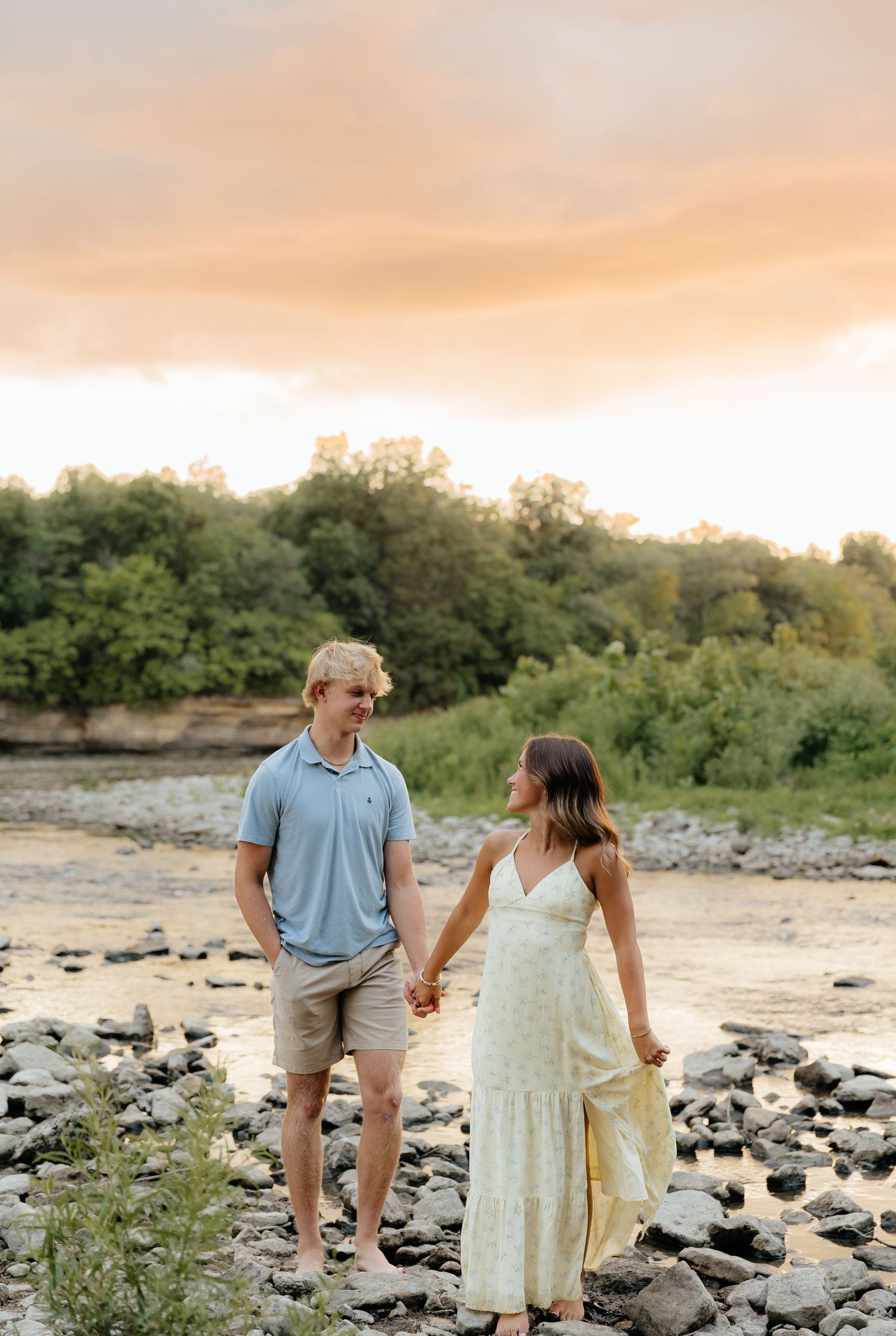 Couple holding hands on rocky riverbank during sunset.