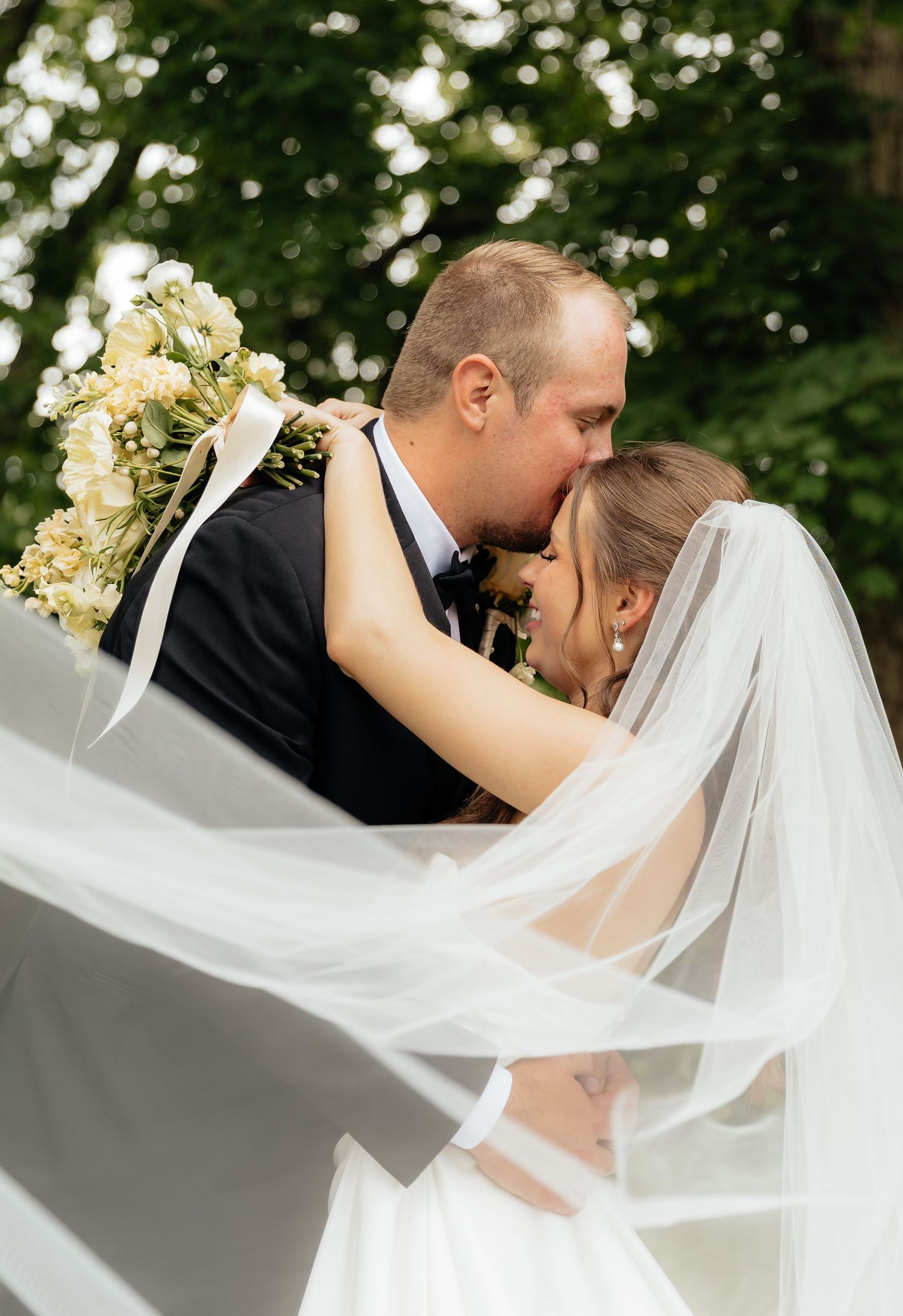 Romantic wedding kiss captured with flowing veil against green foliage backdrop.