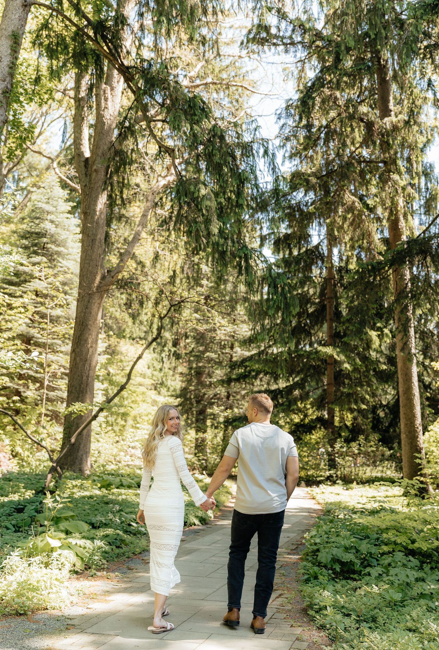 Couple holding hands walking on a forest path surrounded by tall evergreen trees in soft natural light.