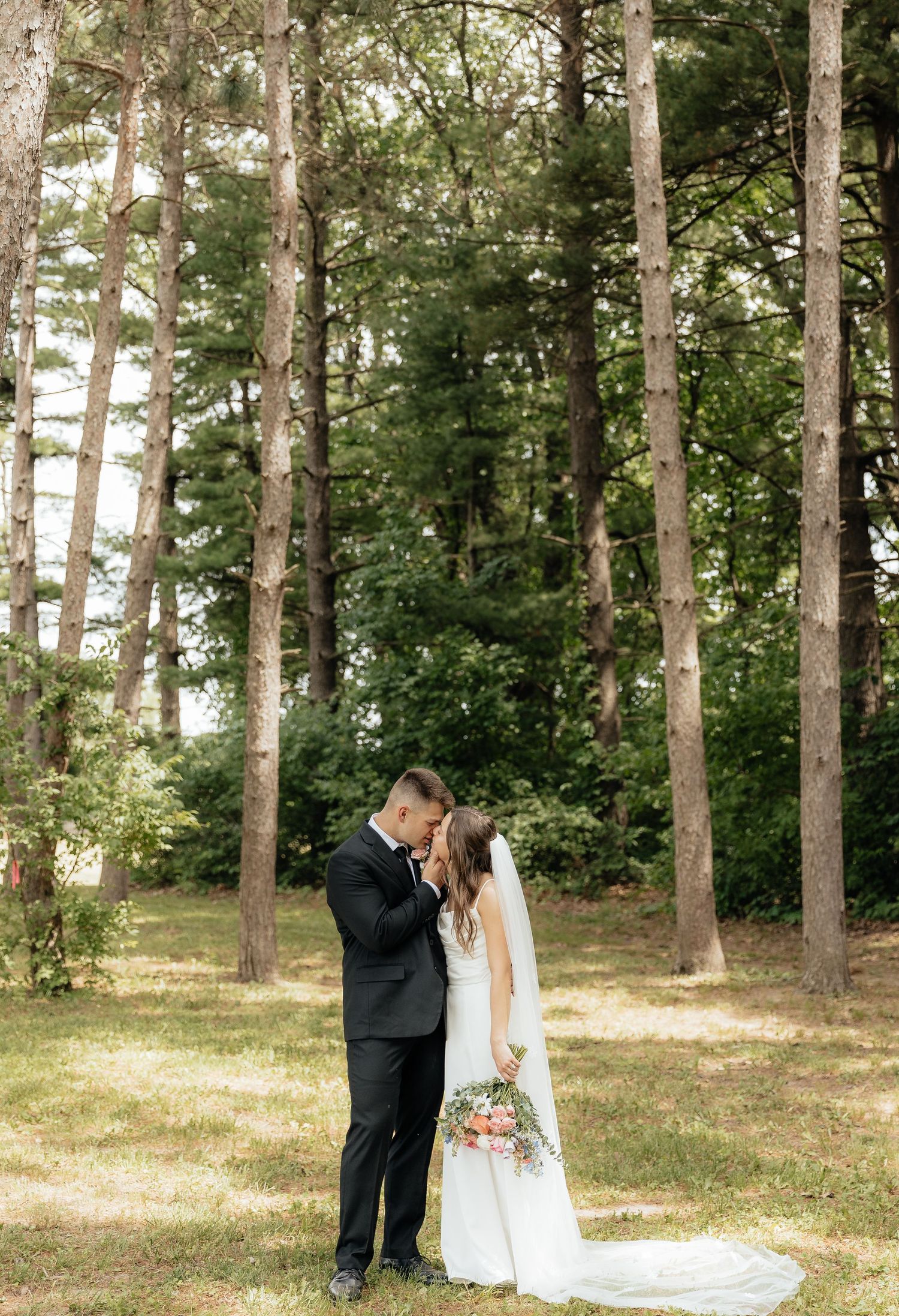 Bride and groom share an intimate moment in a sunlit forest clearing, bride holding a pink floral bouquet.