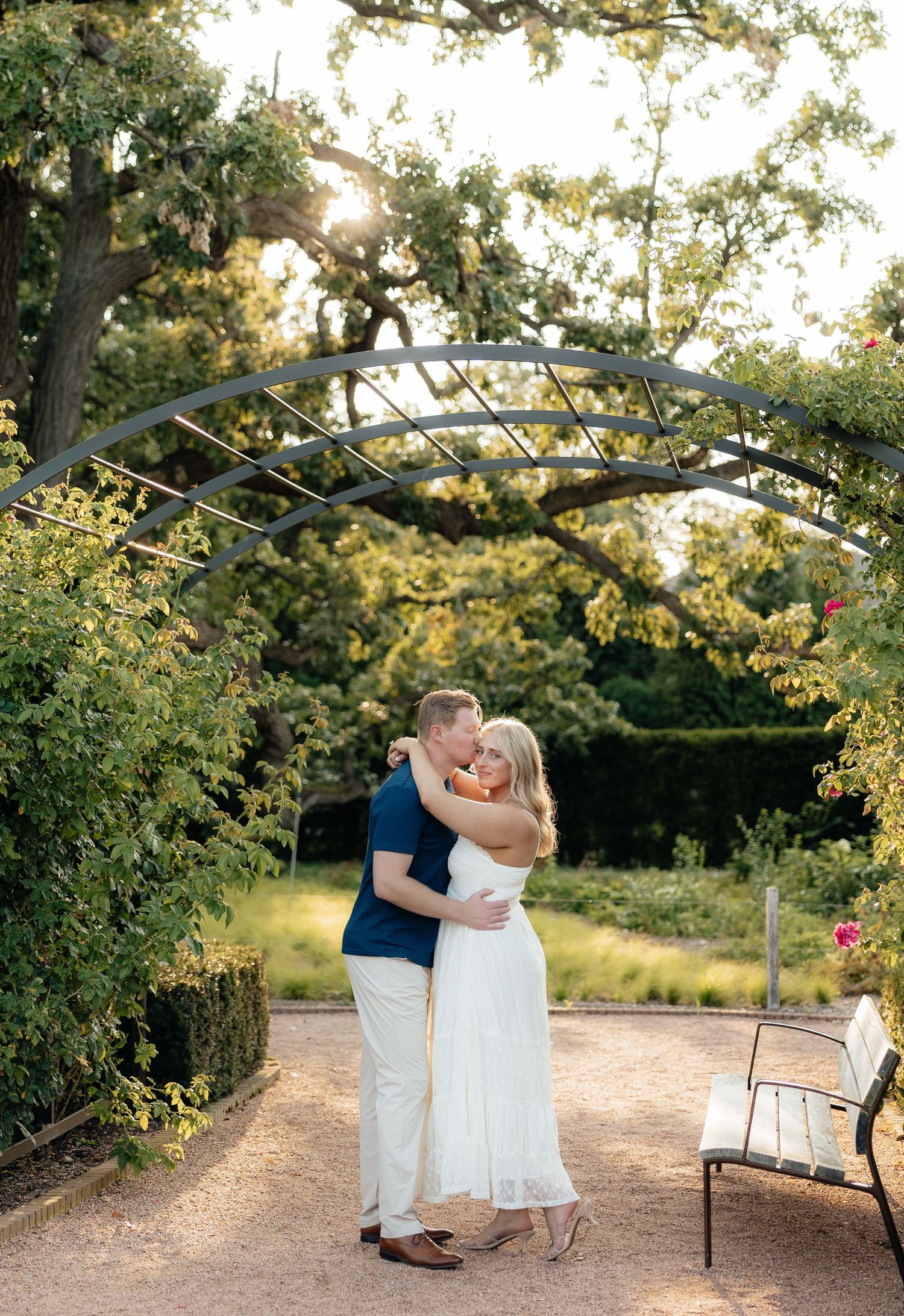 Couple sharing a romantic kiss under a garden arch at golden hour, surrounded by lush greenery and roses.