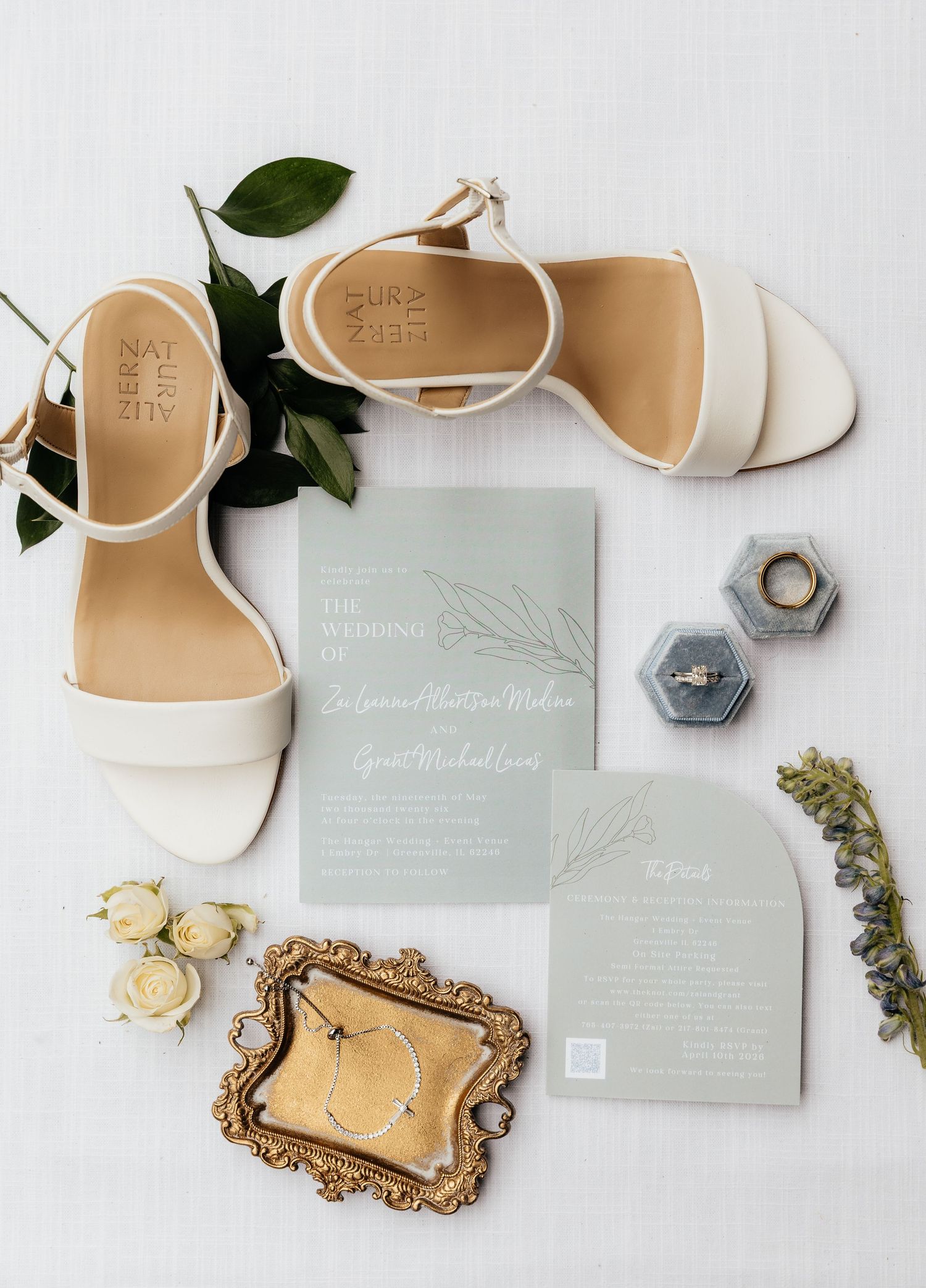 Elegant wedding flat lay featuring white block heel sandals, sage green invitations, ring boxes, pearl earrings, and cream roses.