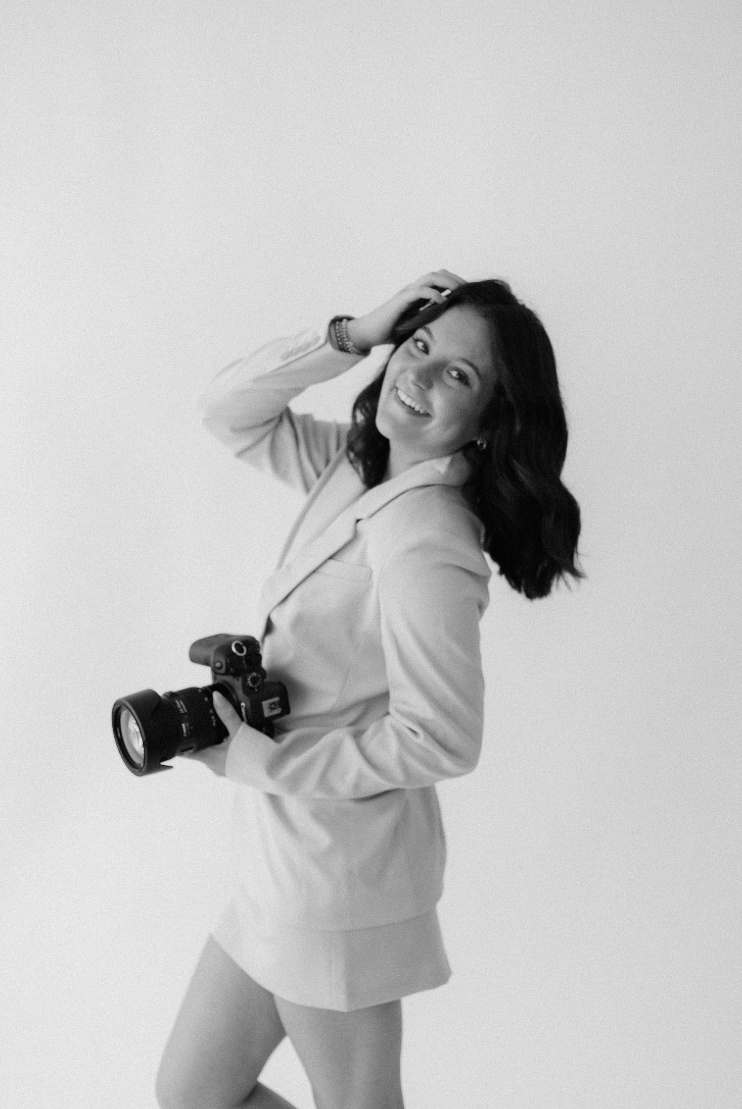 Black and white portrait of a photographer in a light blazer holding a professional camera against a plain background.