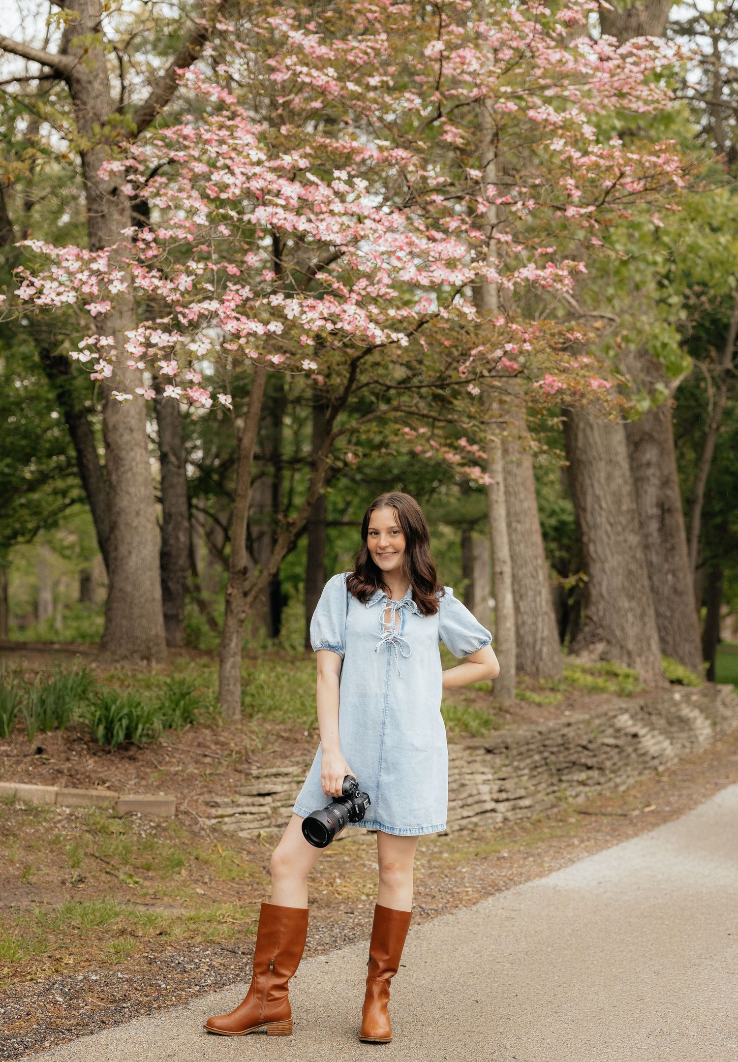 Woman in light blue dress and brown boots holds camera under pink dogwood trees on a scenic path in spring.