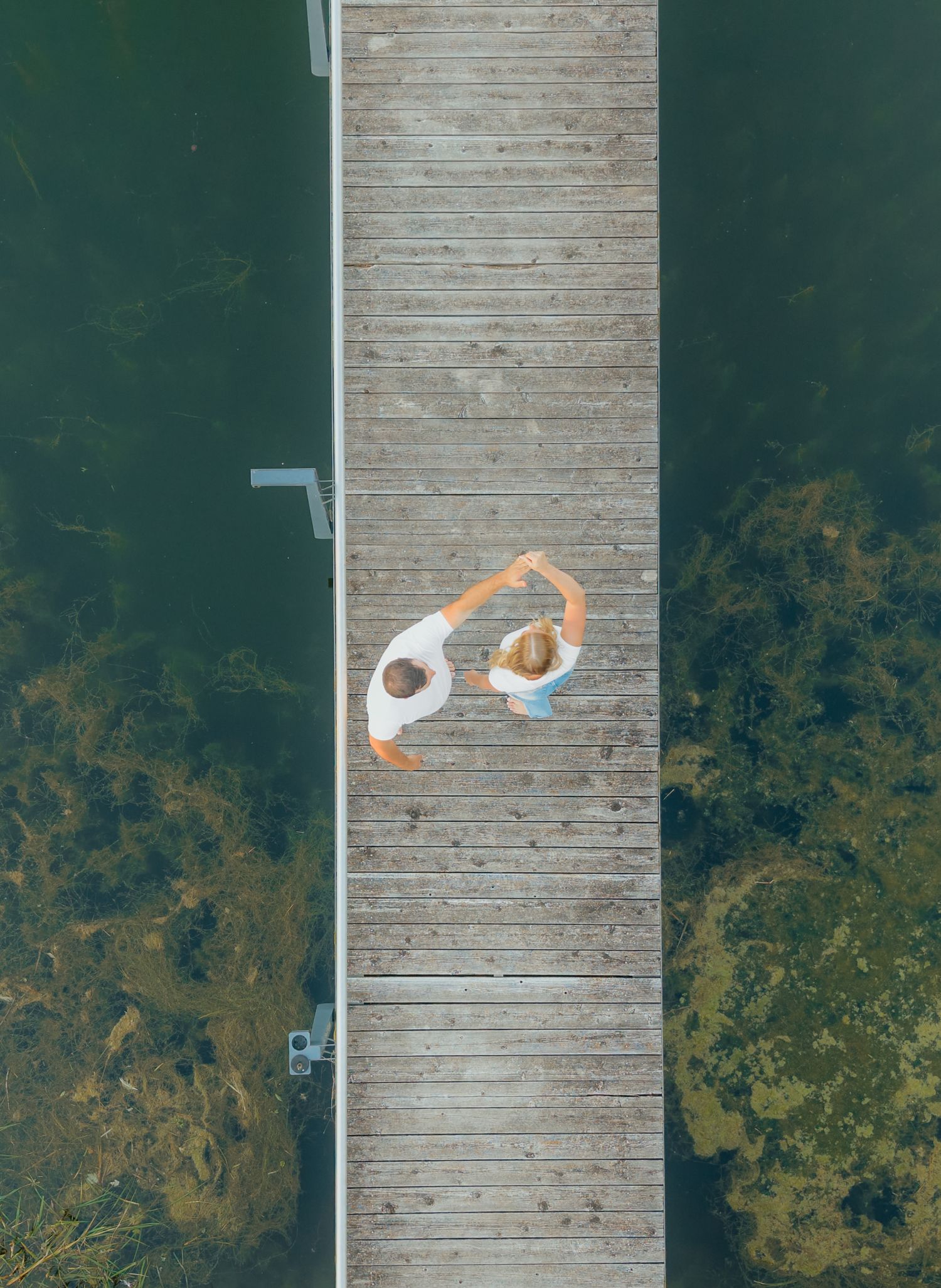 Aerial view of two people forming a heart shape with their arms on a wooden dock surrounded by clear green water.