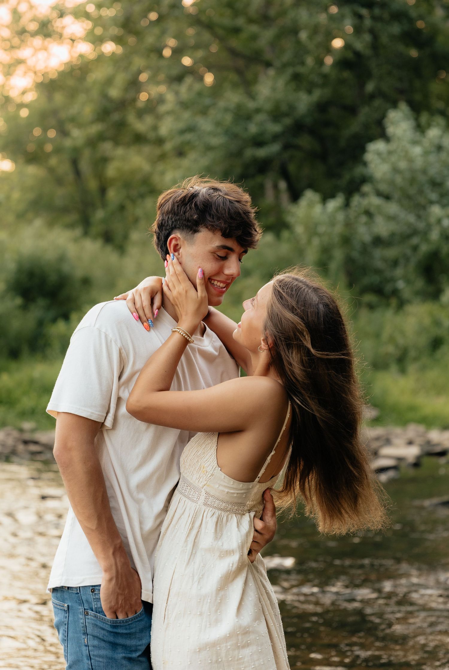 A couple shares an intimate moment along a riverbank at sunset with trees and soft golden light in the background.