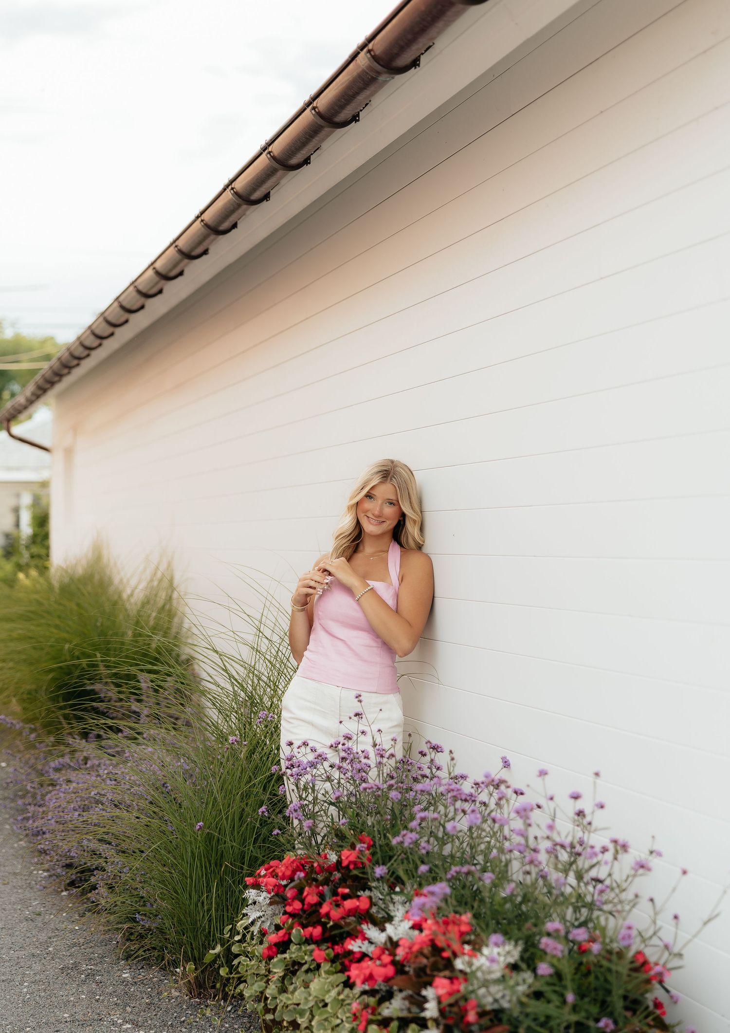 Smiling blonde woman in pink top leaning against white wall surrounded by colorful garden flowers.