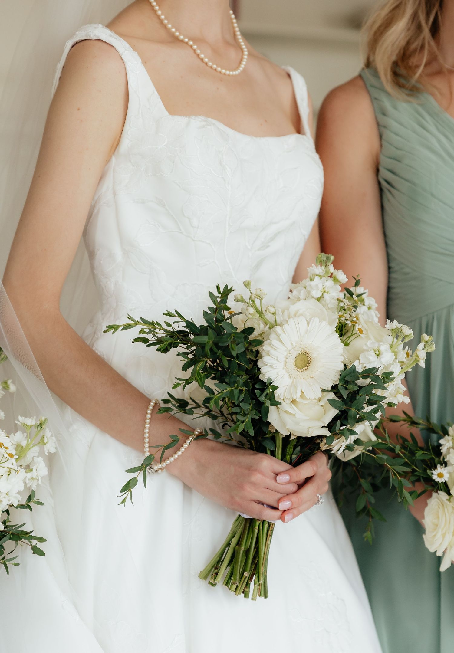 Bride in white gown holding white floral bouquet with greenery, wearing pearl necklace, standing beside sage green bridesmaid.
