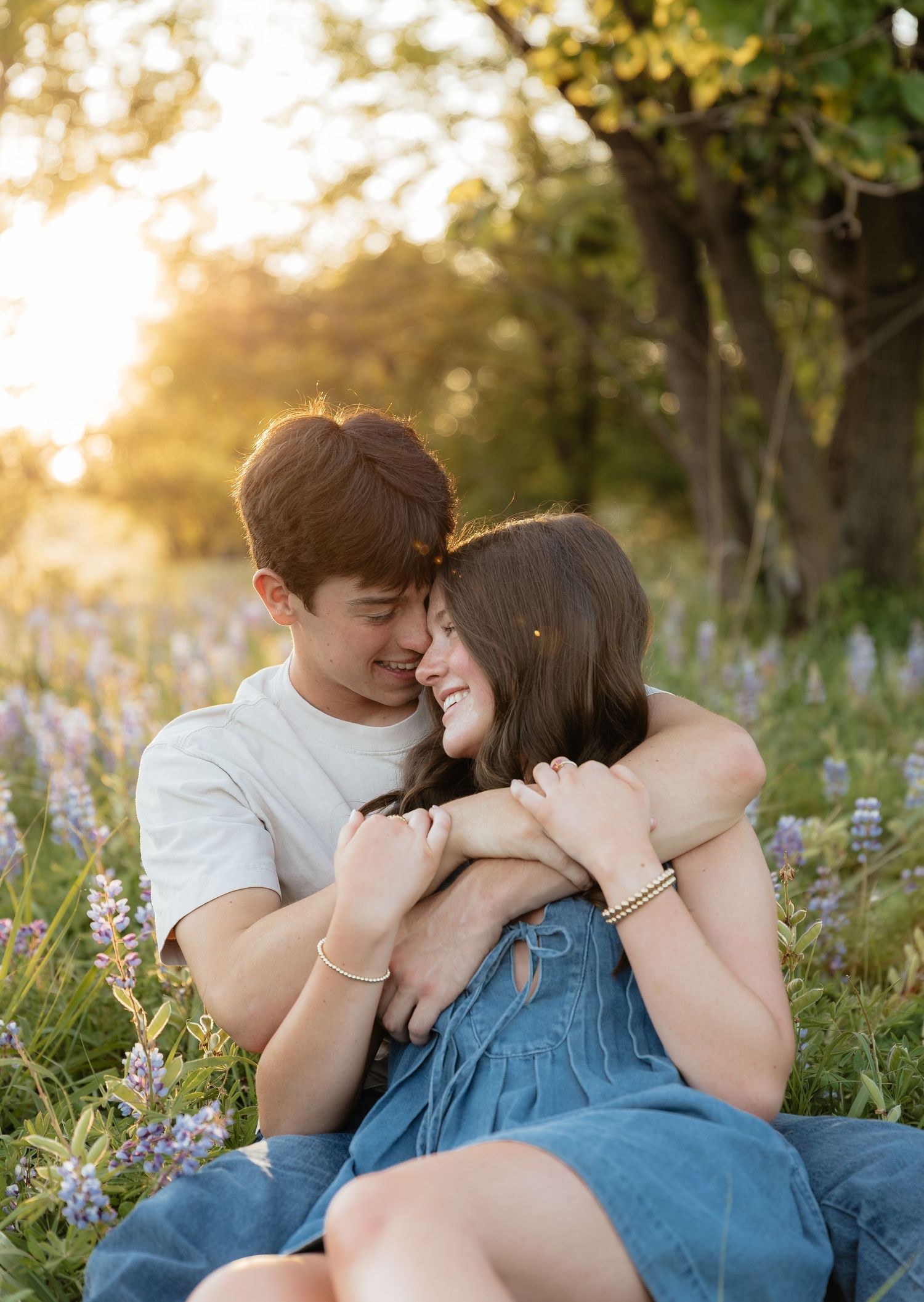 A couple sits in a wildflower field at golden hour, smiling and embracing romantically.