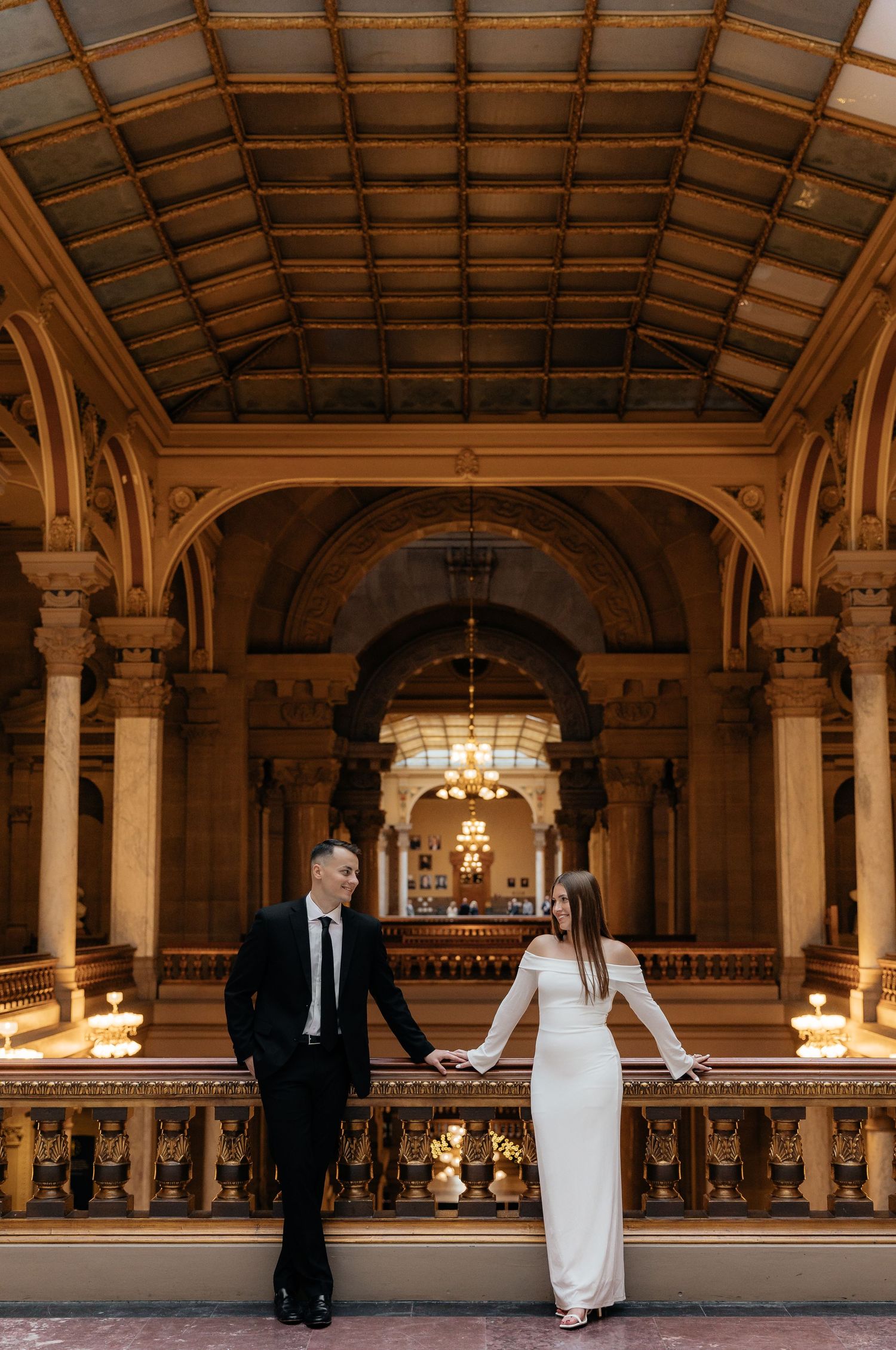 Elegant couple in formal attire poses in grand historic ballroom with ornate architecture and chandeliers.