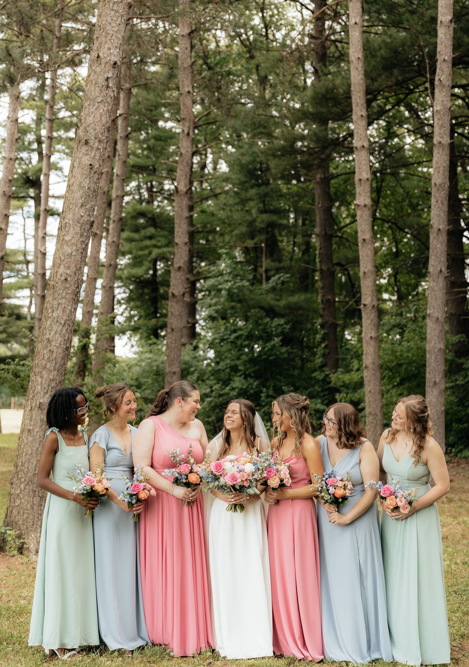Bridesmaids in mismatched pink and mint gowns laugh together holding colorful bouquets among tall pine trees.