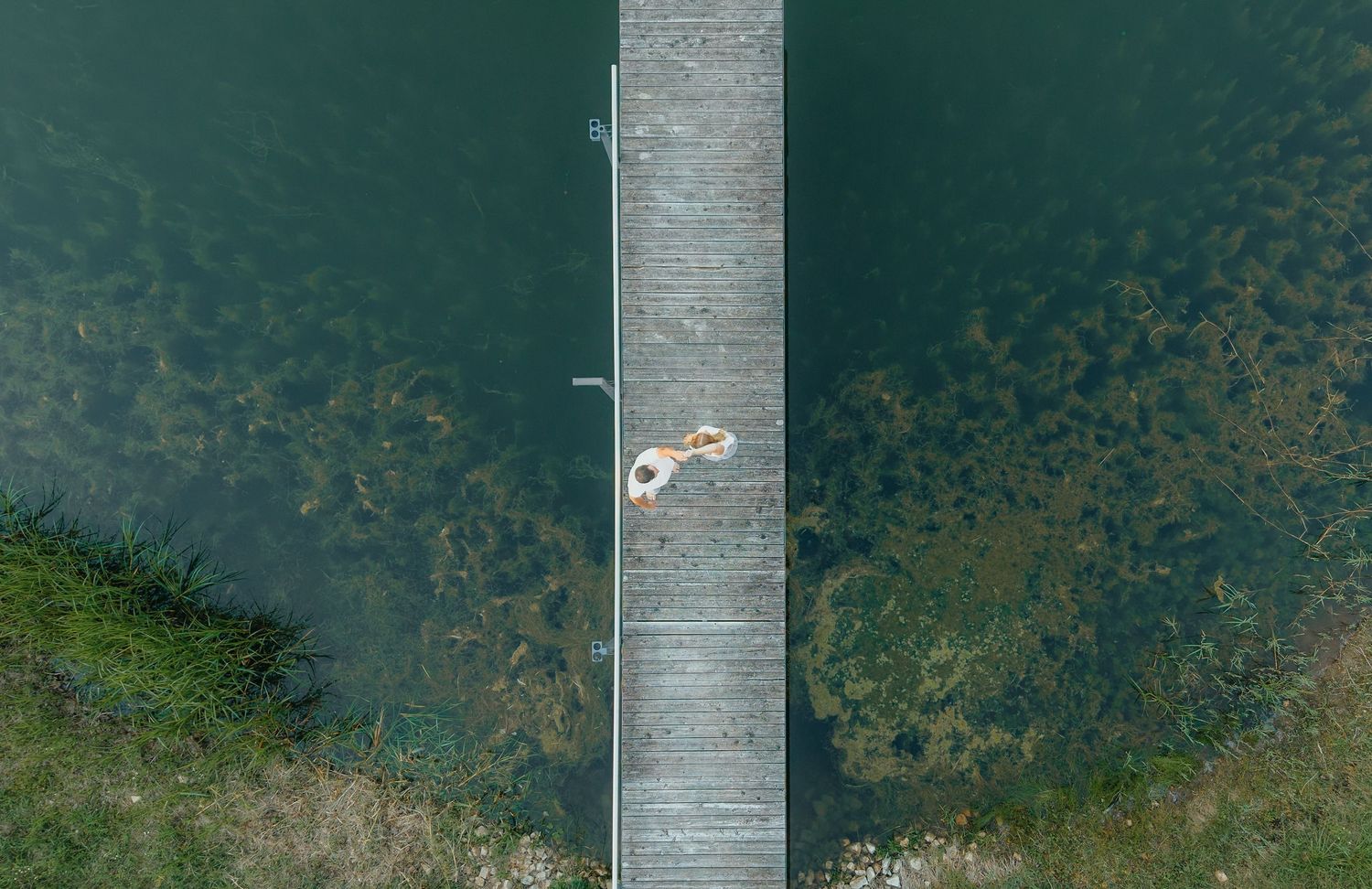 Aerial view of a person lying on a wooden dock pier extending over calm green lake water.