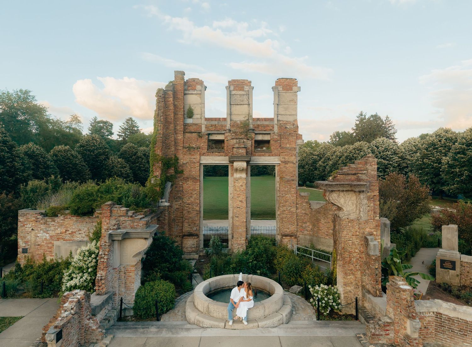 Couple embracing before historic brick ruins with lush greenery at golden hour.