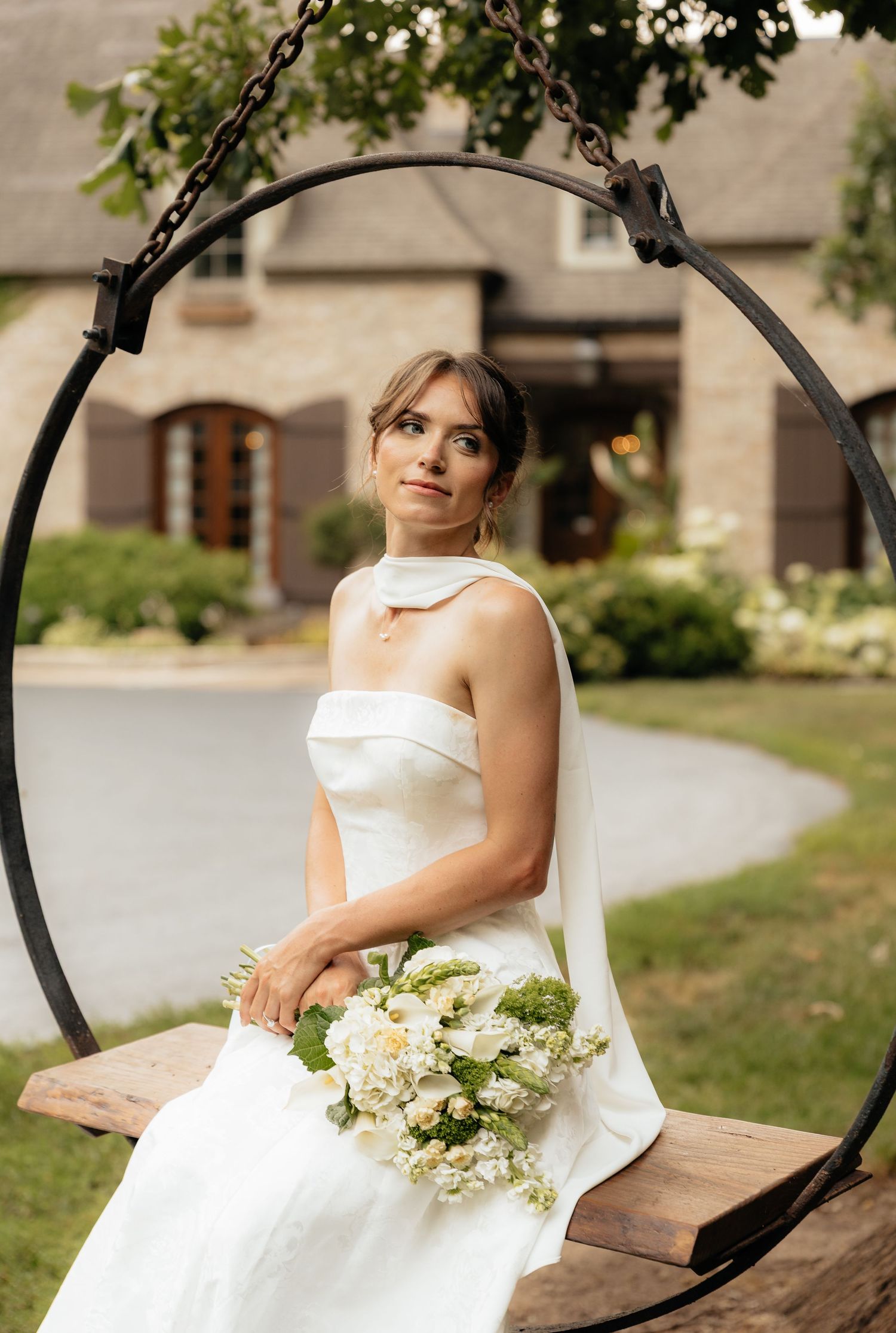 Bride sitting on wooden swing holding white floral bouquet in garden setting.