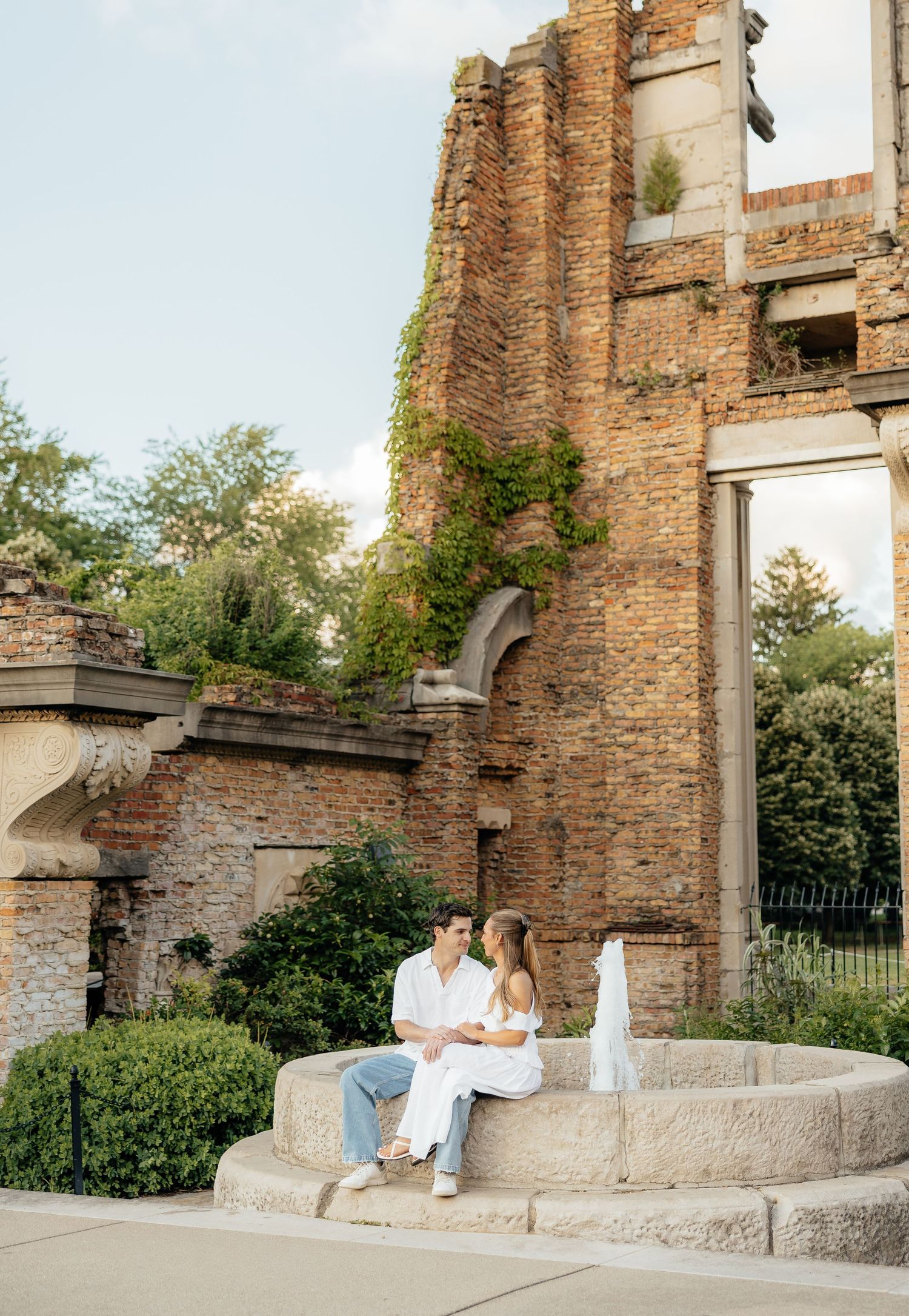 Couple sitting on a stone fountain near a rustic brick ruin covered in ivy, surrounded by lush greenery.